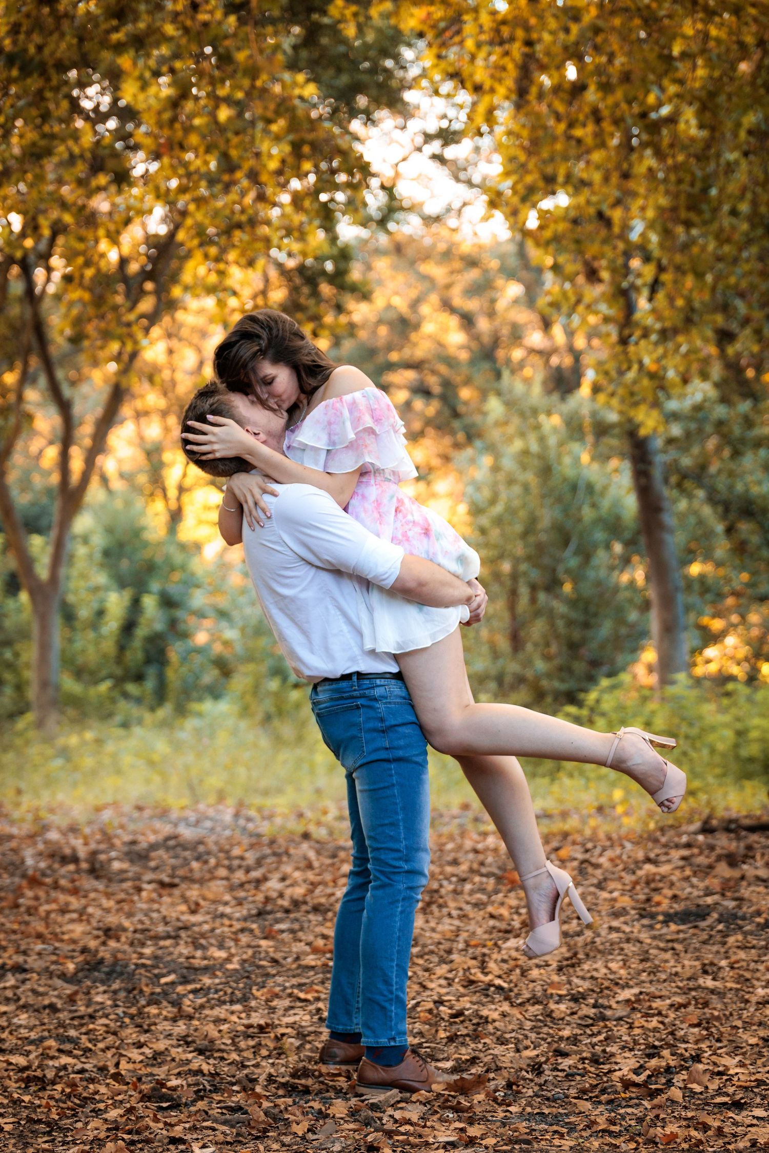 A romantic couple embraces in a golden autumn forest setting with fallen leaves scattered on the ground.