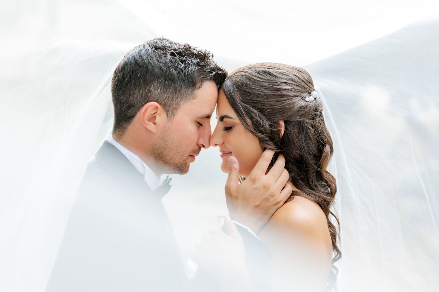 Romantic bride and groom touching foreheads under flowing white veil in an intimate wedding portrait.