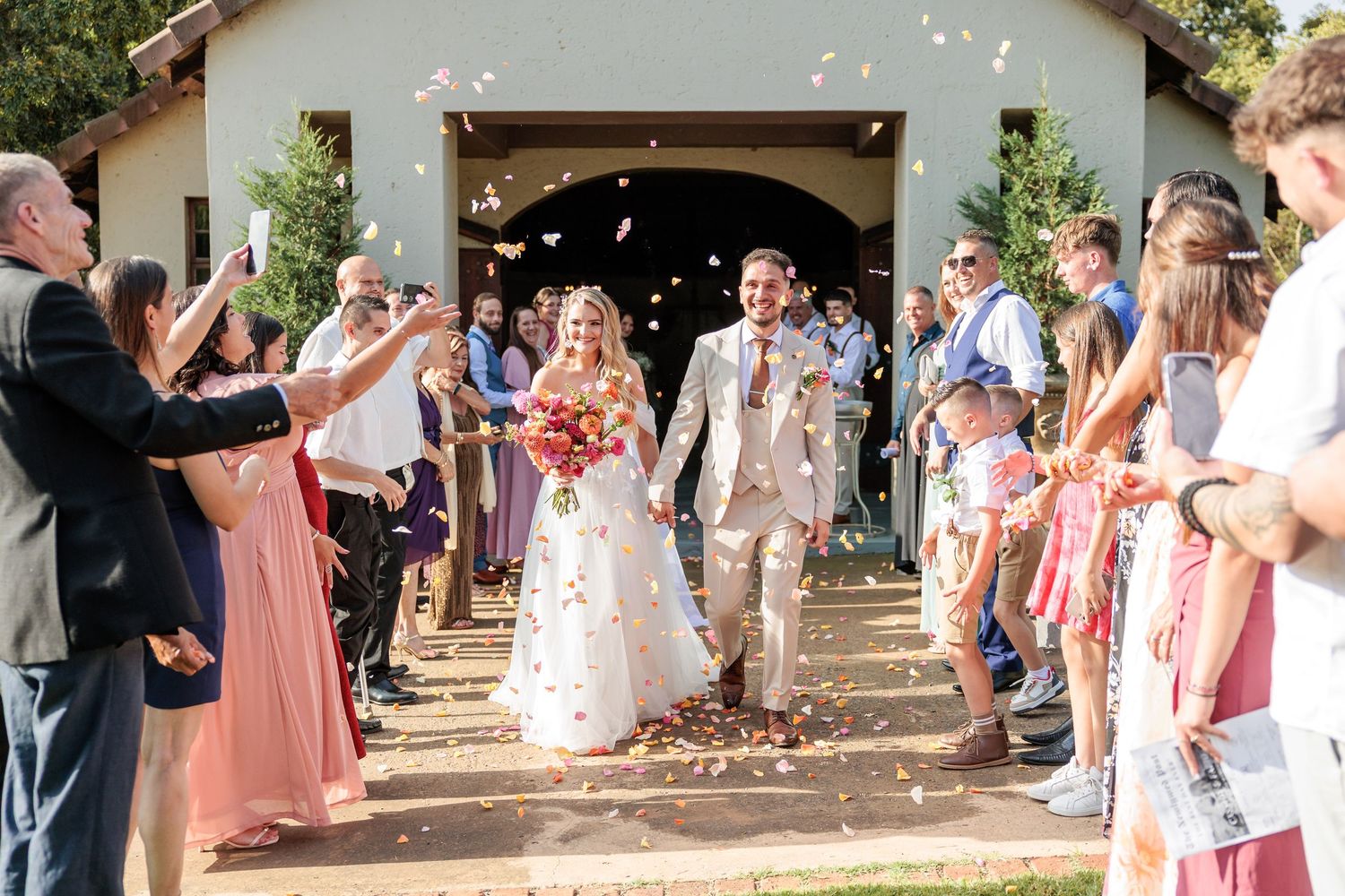 Newlywed couple walks through confetti shower as guests celebrate outside a rustic white chapel wedding venue.