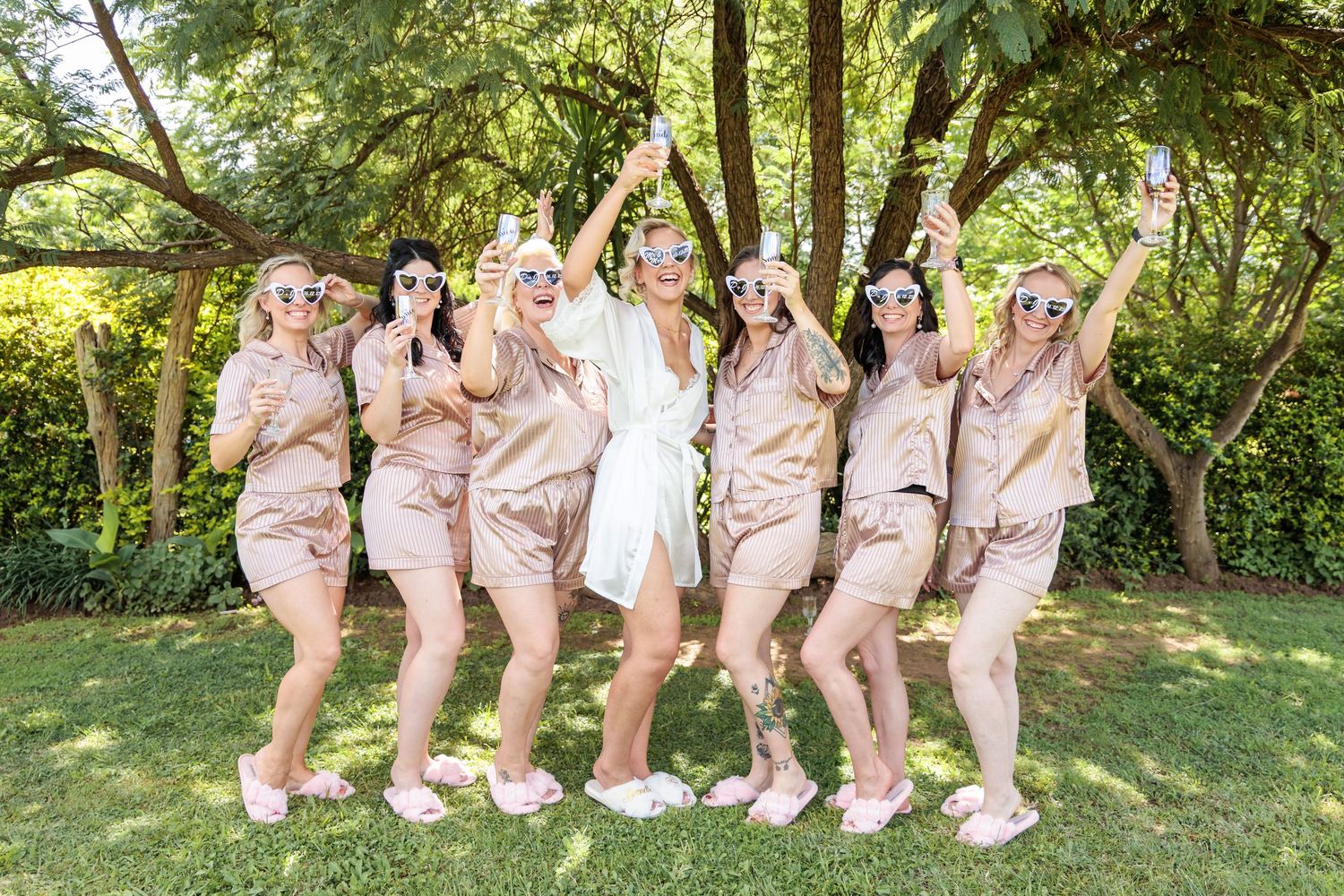 A wedding party in blush pink dresses poses together outdoors in a garden setting during a summer celebration.