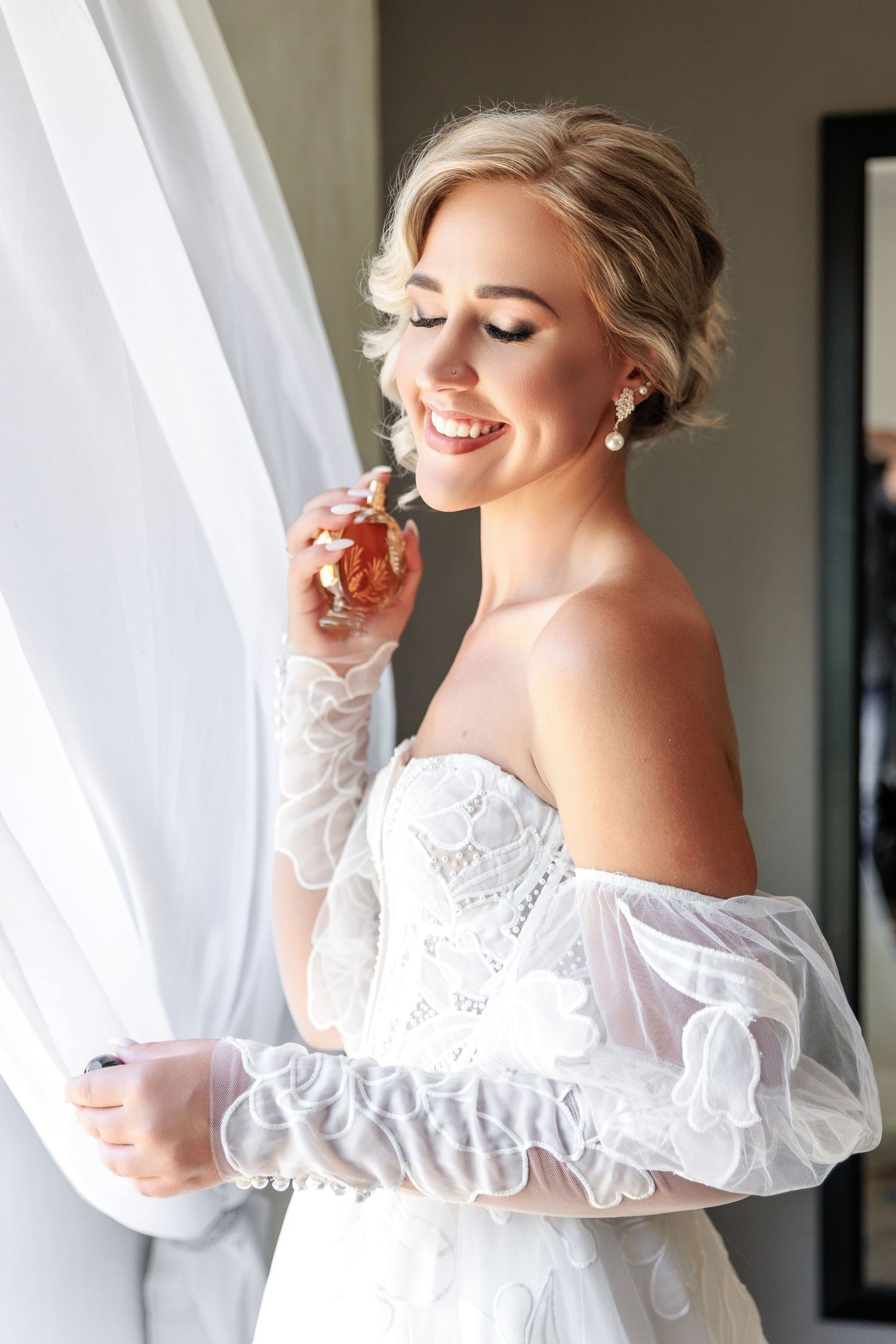 A bride in an elegant off-shoulder white lace gown smiles while standing by a window in soft natural light.
