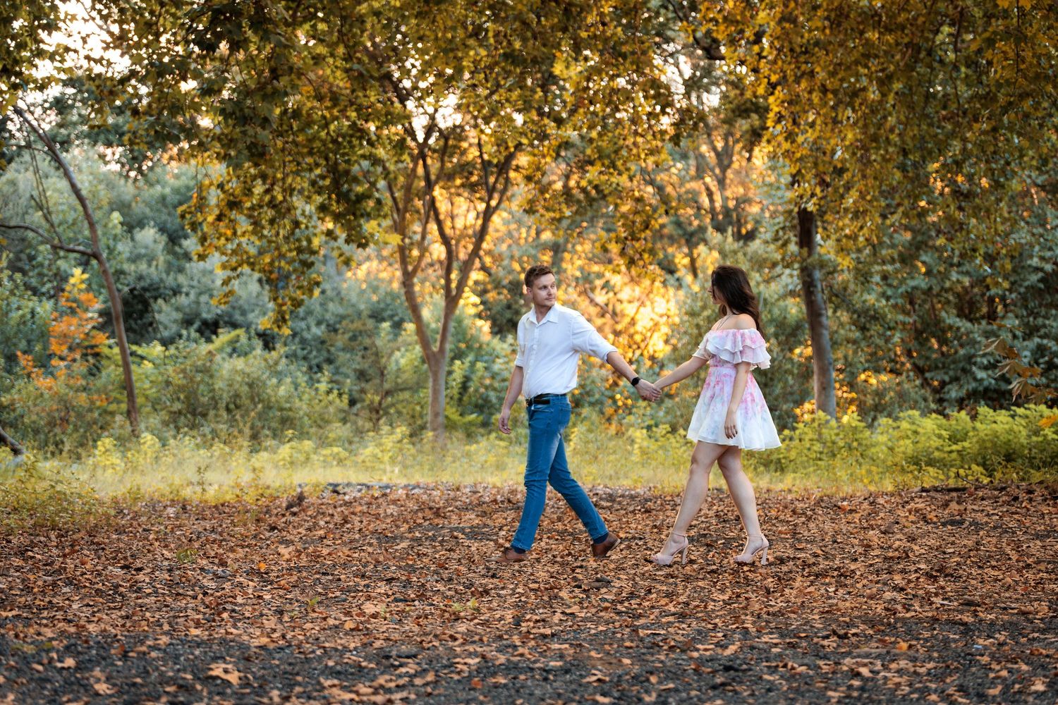 A couple walks hand in hand through a scenic autumn landscape with golden leaves and sunlight filtering through trees.