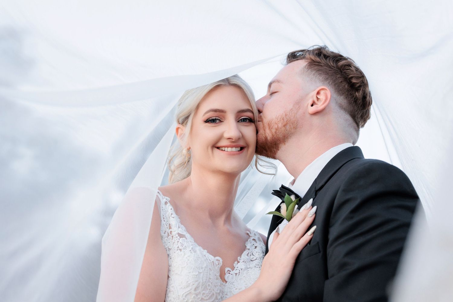 Groom in black tuxedo kissing bride on cheek under flowing white veil on wedding day.