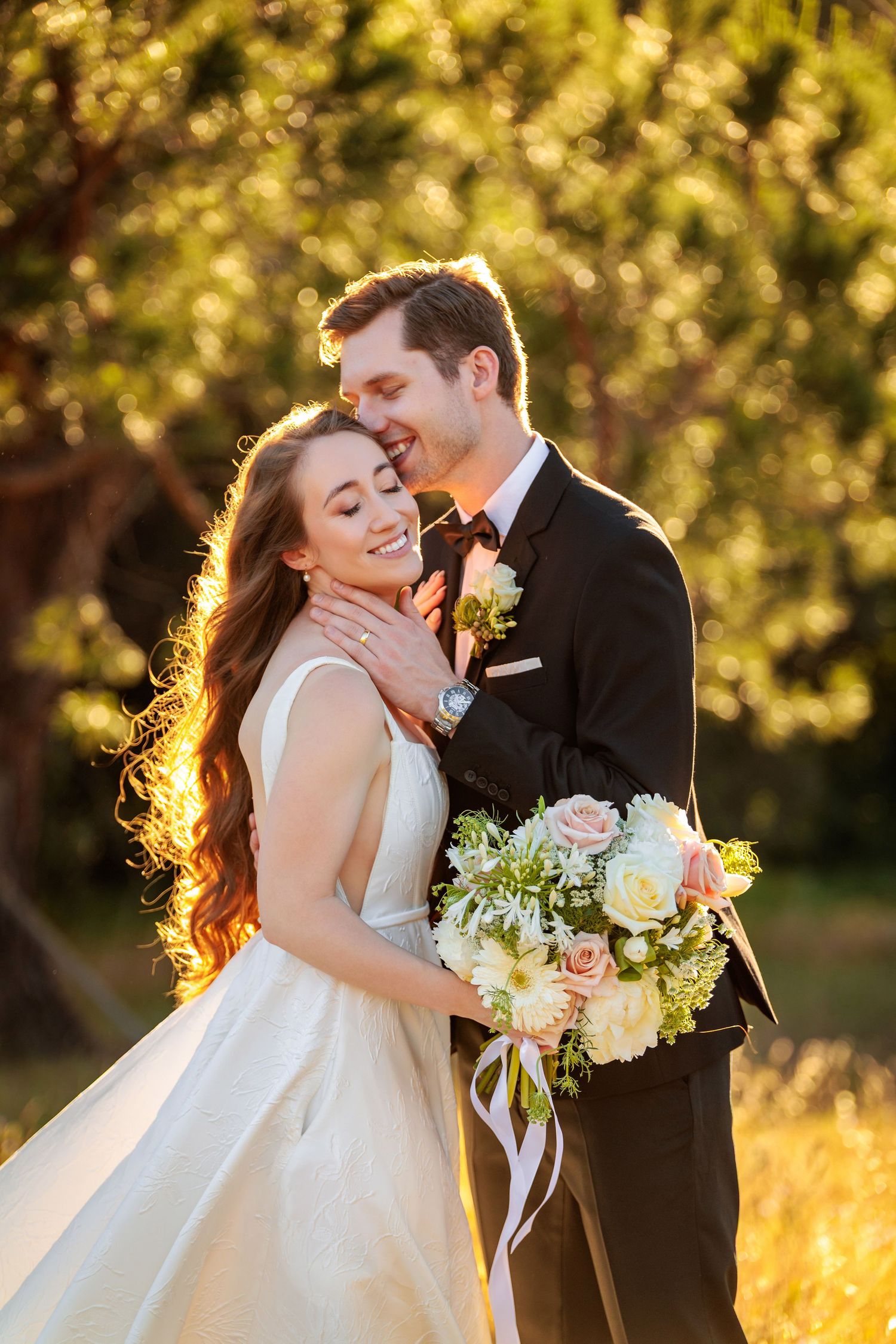 Bride and groom embrace outdoors in golden sunlight, she holds a white and pink bouquet while wearing a white gown.
