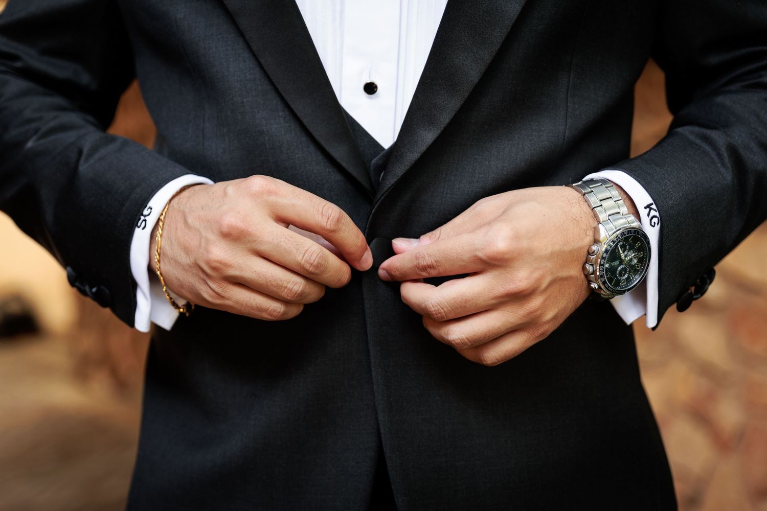 Man in black tuxedo buttoning jacket, wearing silver watch and cufflinks, formal wedding attire close-up.