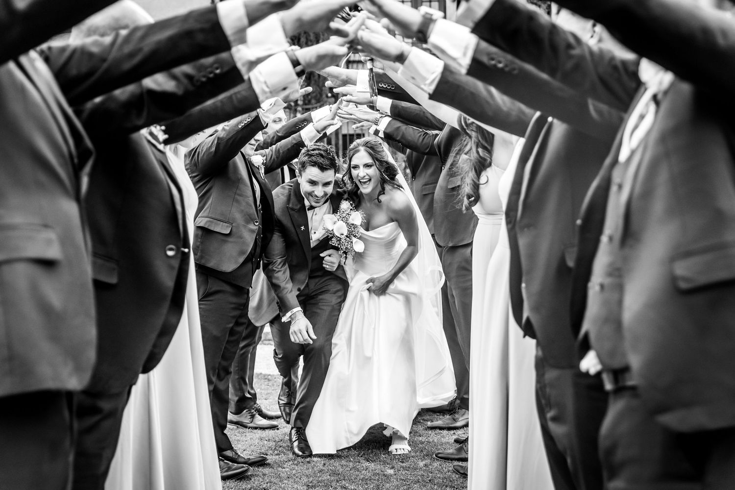 Black and white photo of a bride and groom walking through an arch of raised hands formed by their wedding party.