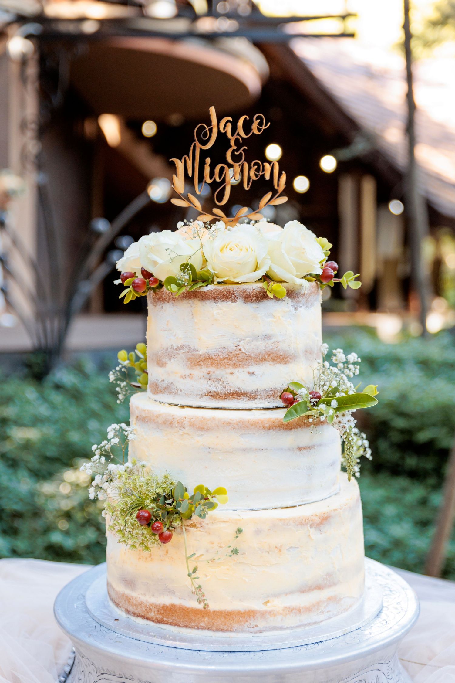 Semi-naked wedding cake decorated with fresh greenery and gold topper on white stand.