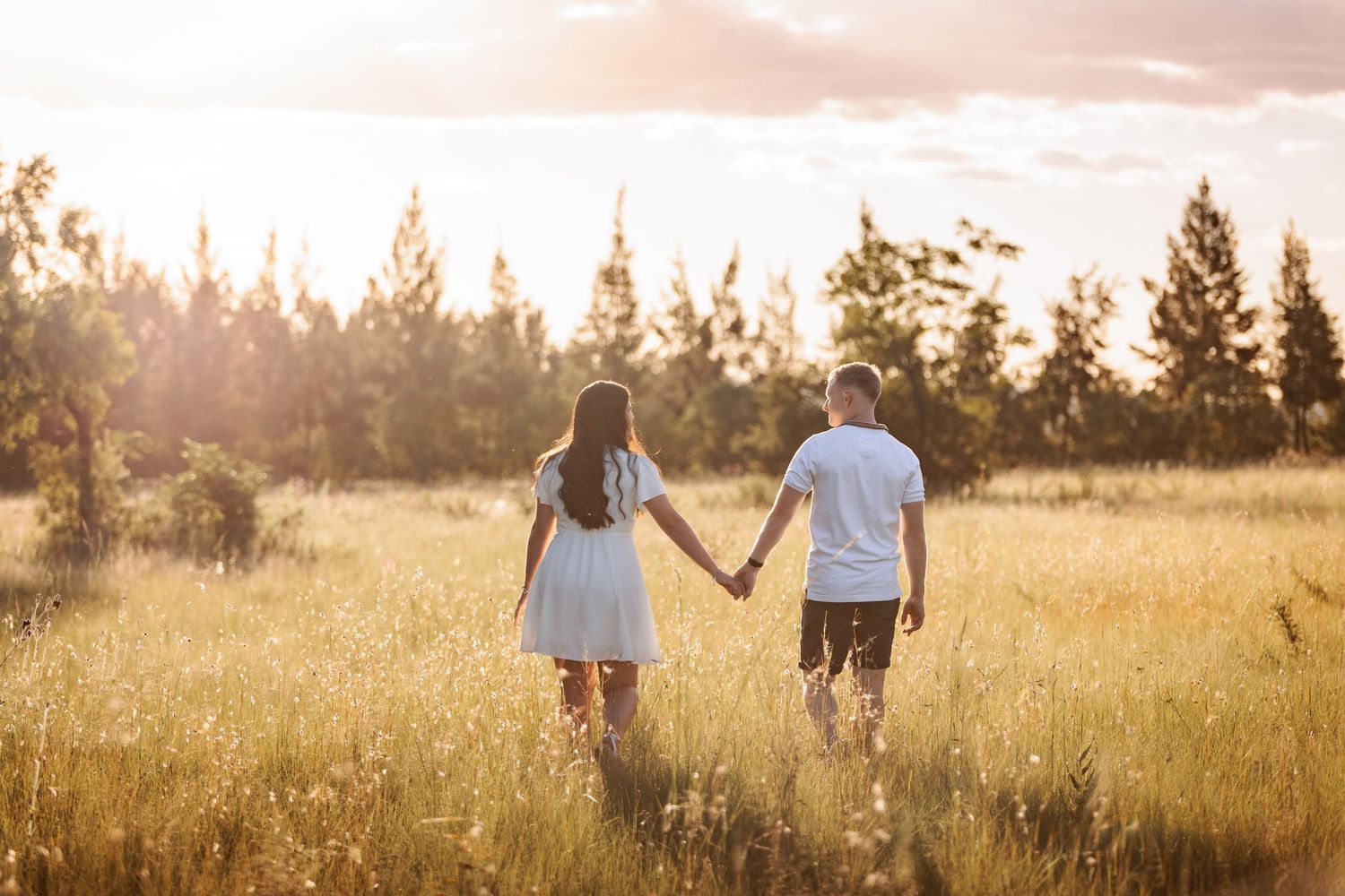 A couple walks hand in hand through a golden sunlit meadow at sunset with evergreen trees in the background.