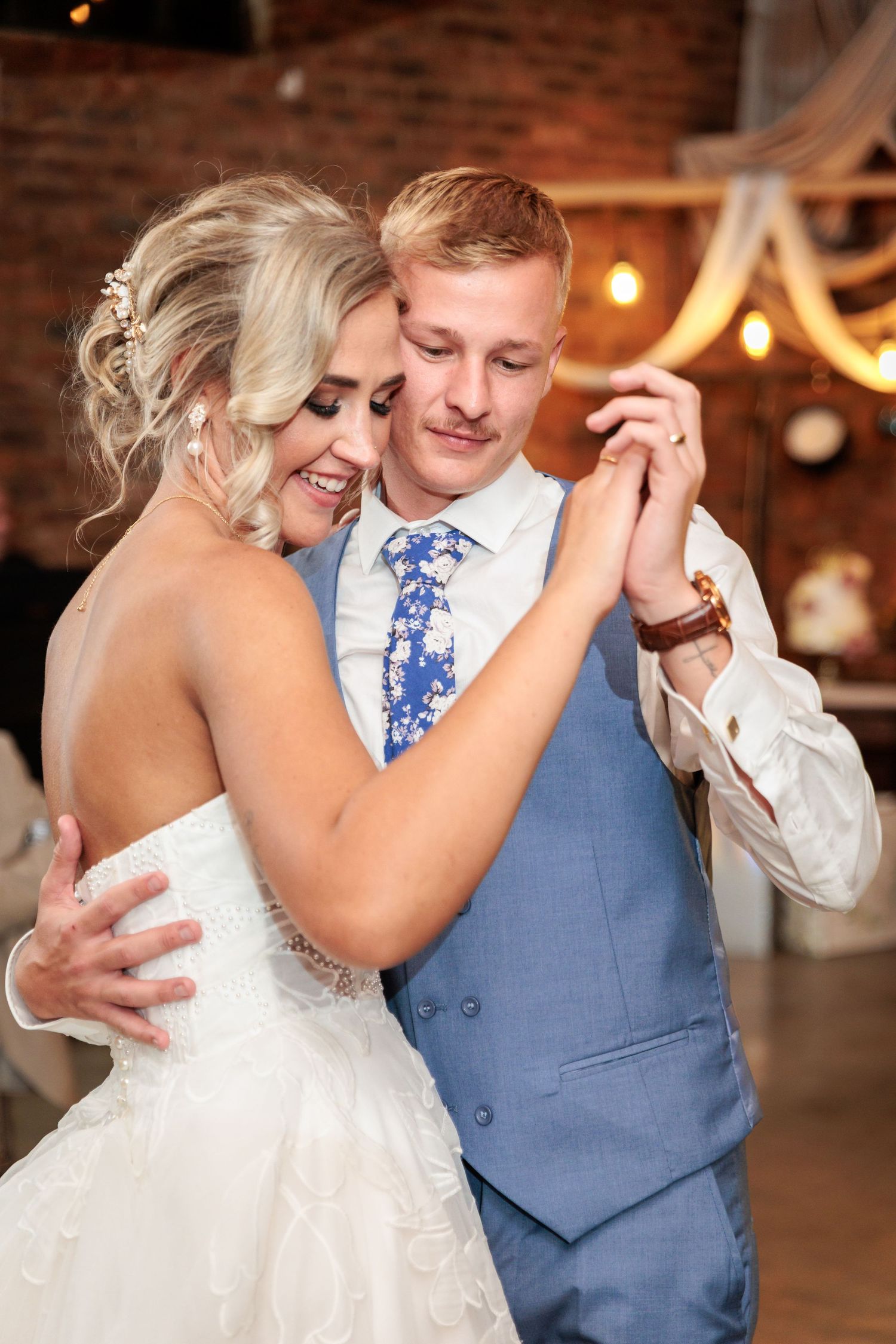 A couple shares an intimate dance moment at their wedding reception in a rustic brick-walled venue with twinkling lights.