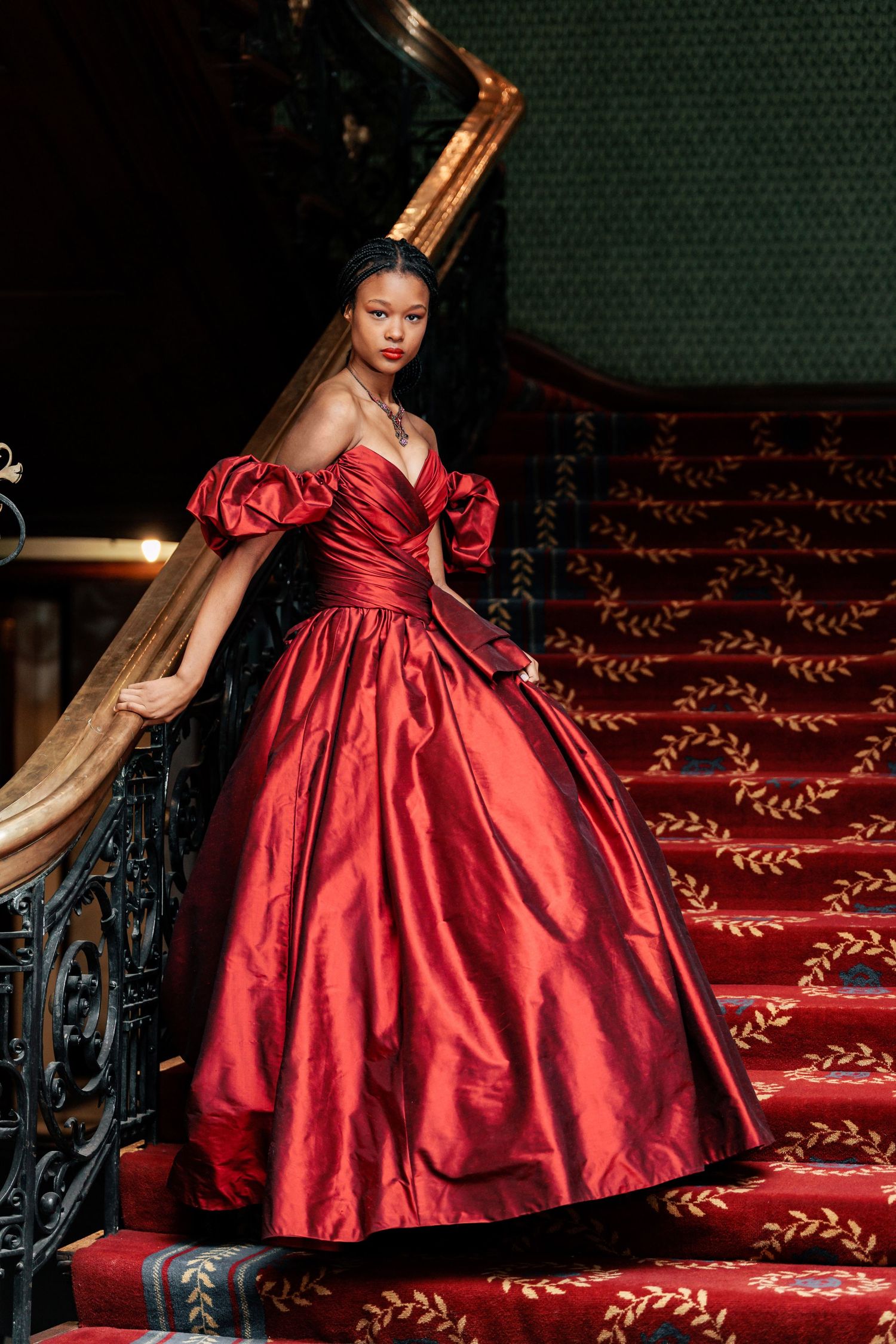 Woman in luxurious red satin ball gown poses gracefully on ornate carpeted staircase with decorative metal railing.
