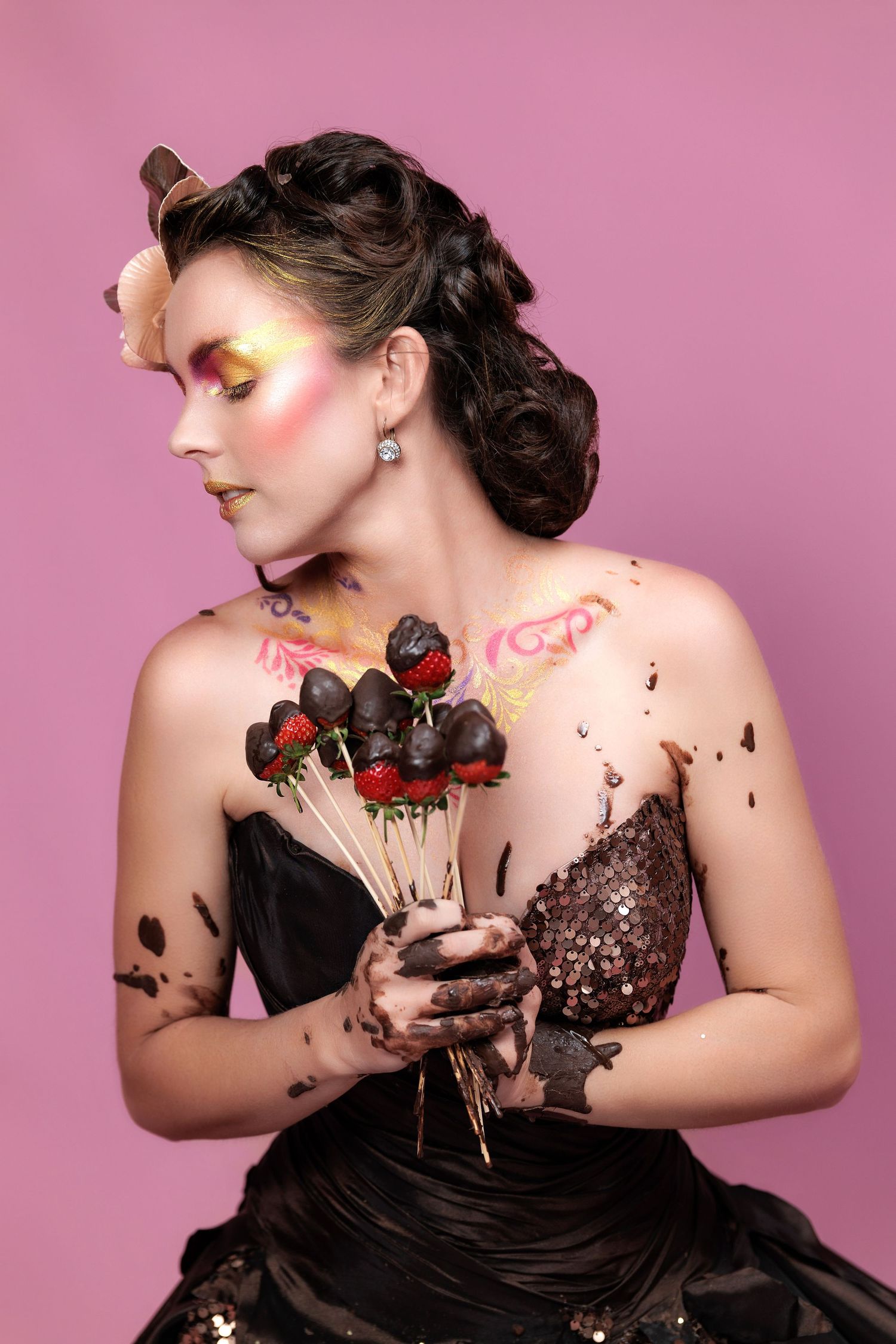 Artistic fashion portrait with chocolate covered strawberries against pink backdrop featuring dark gown and dramatic makeup.