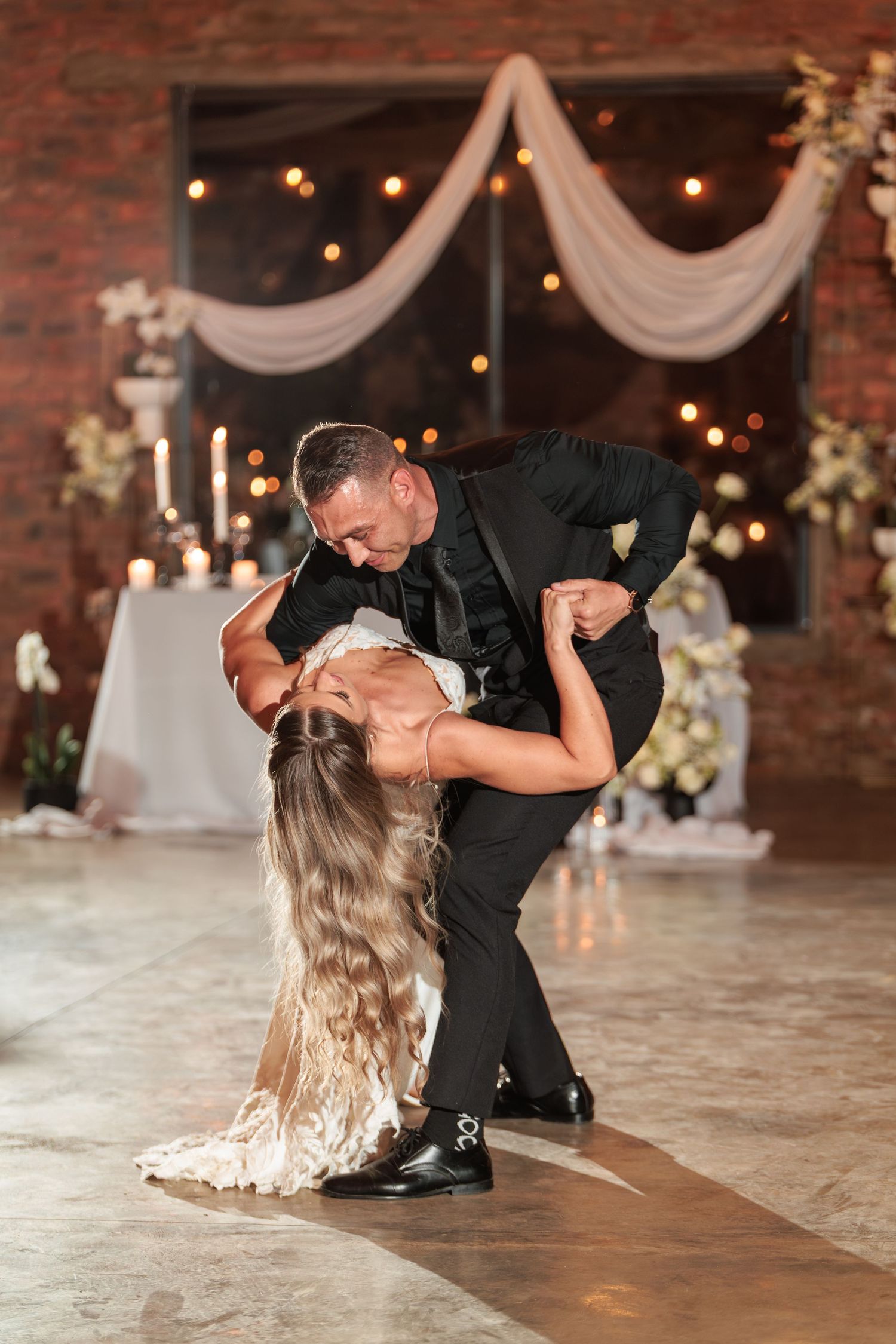 A couple performs a romantic dip dance move at their wedding reception with string lights and white draping in background.