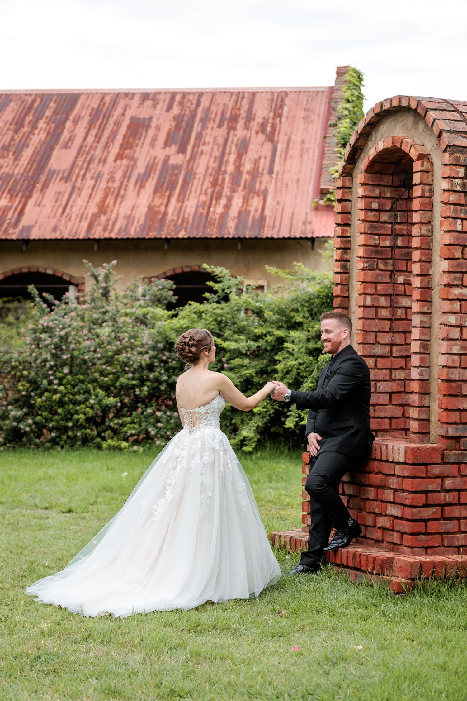 A couple in wedding attire shares a sweet moment by a rustic brick structure in a garden setting.