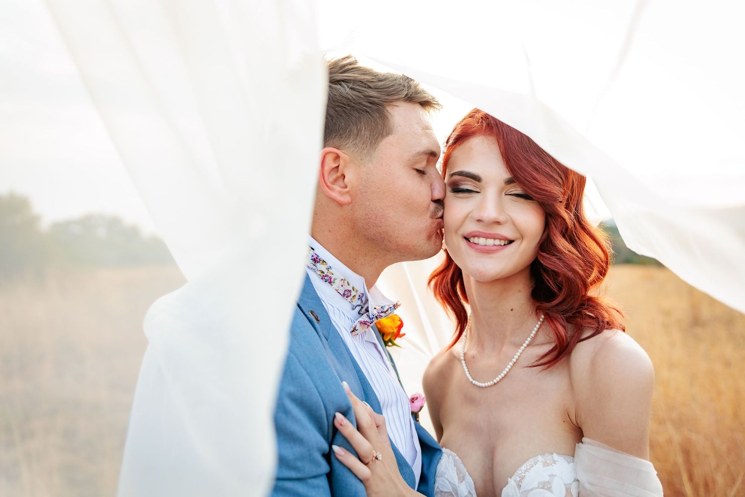 A couple shares a tender moment in a sunlit field, with the bride wearing a white gown and pearl necklace.