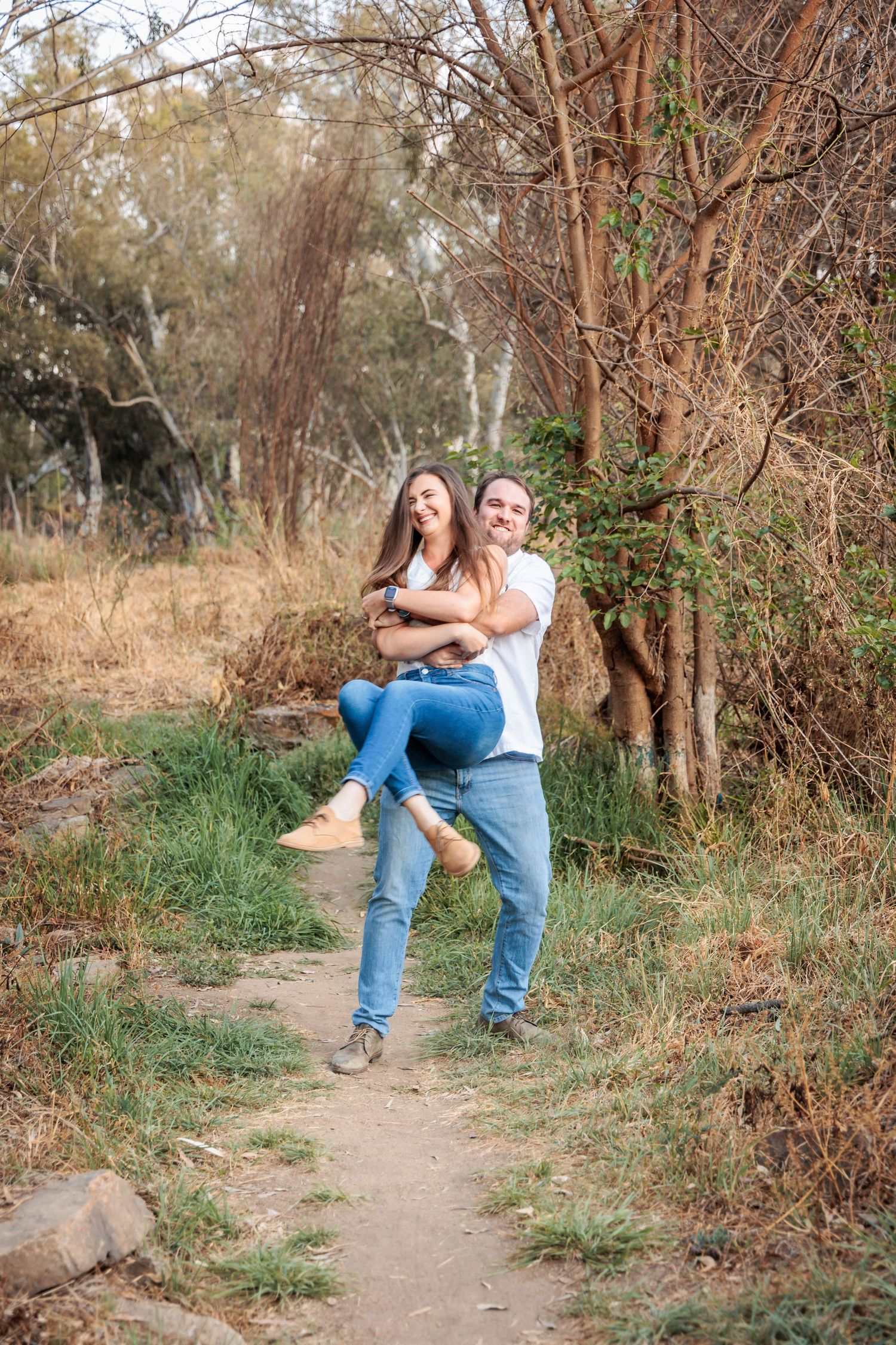 A couple shares a playful moment on a dirt path surrounded by autumn trees and brush.