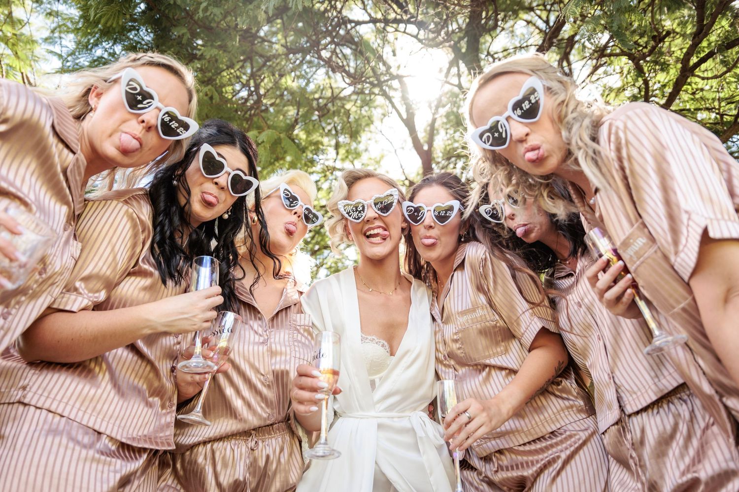 Bride in white surrounded by bridesmaids in striped pajamas wearing heart sunglasses, celebrating outdoors.