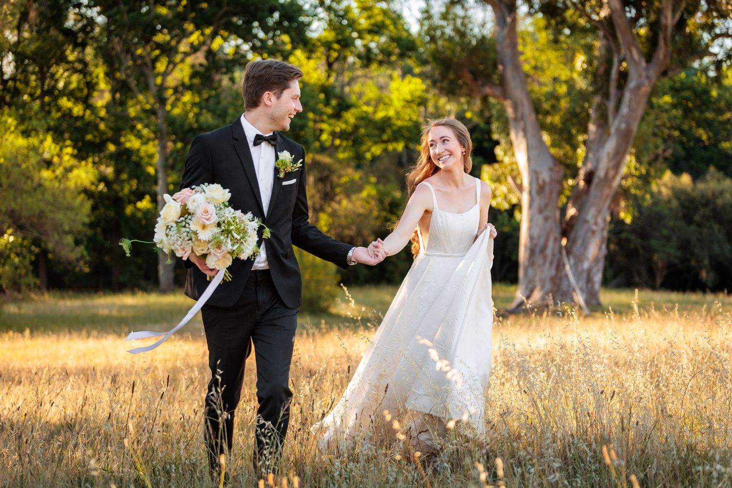 Bride and groom walking hand in hand through golden meadow, bride holding bouquet, groom in tuxedo, trees in background.