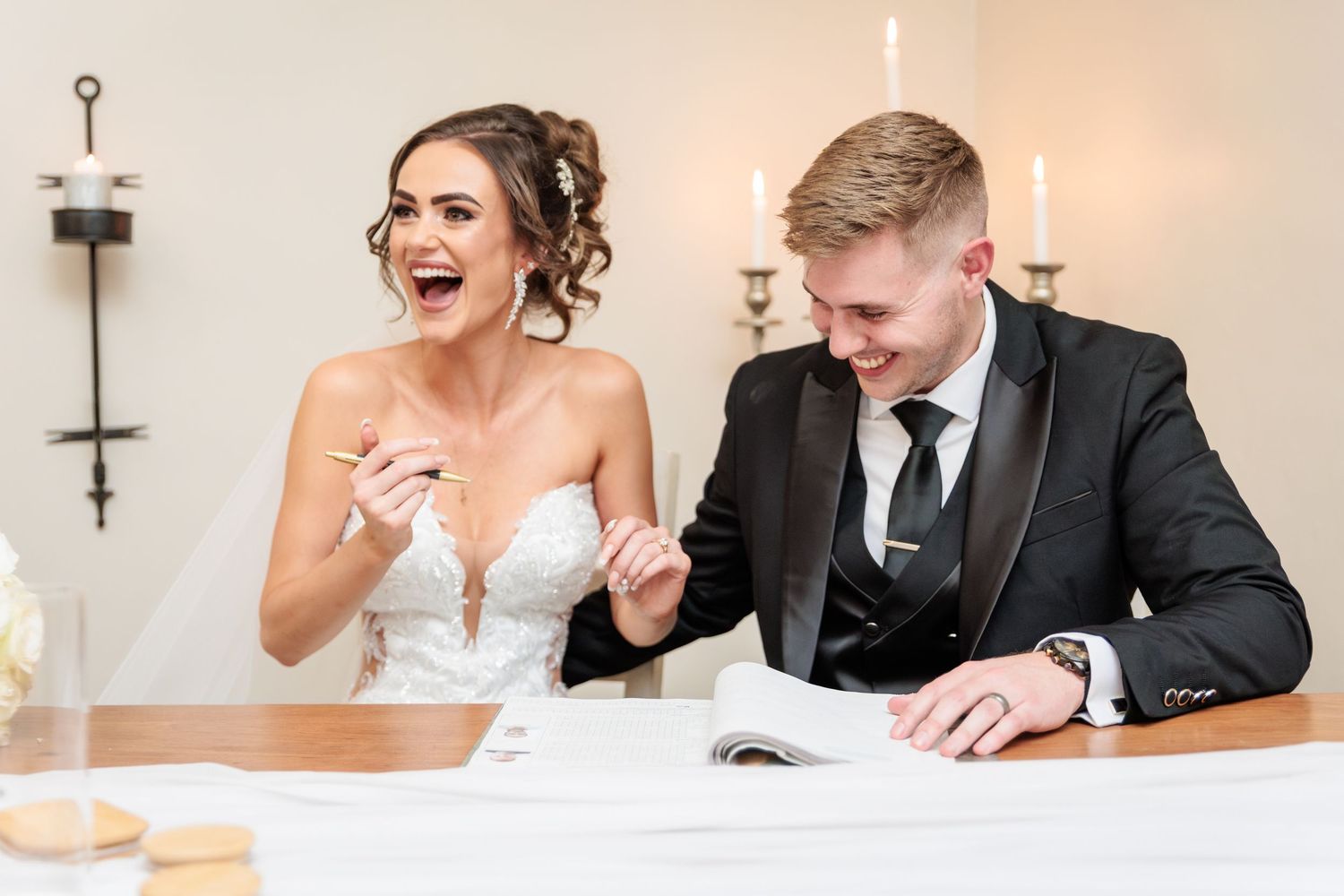 A wedding couple shares a joyful moment laughing together at a reception table with white decor and candles.