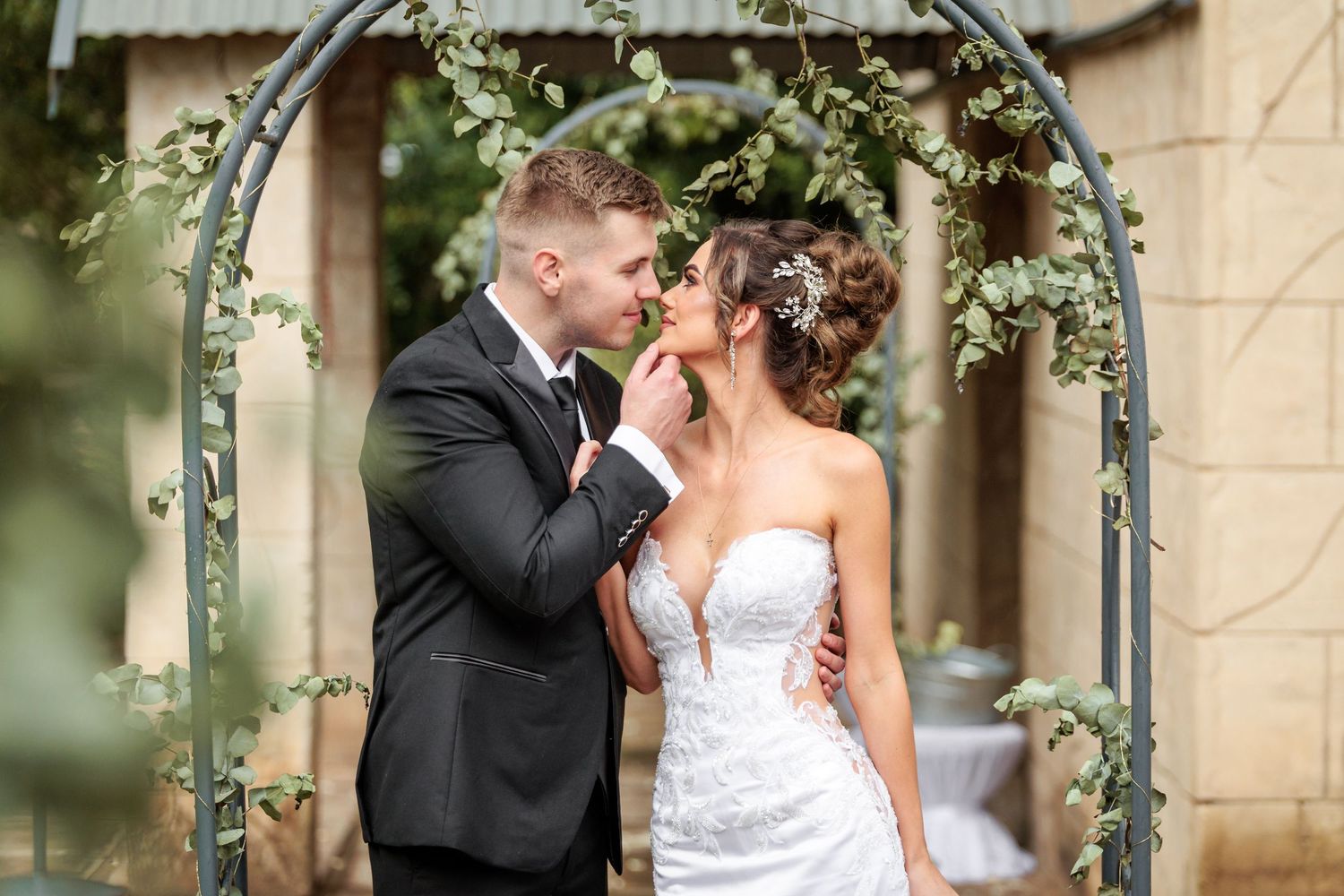 Romantic couple shares tender moment under floral garden arch during outdoor wedding ceremony.