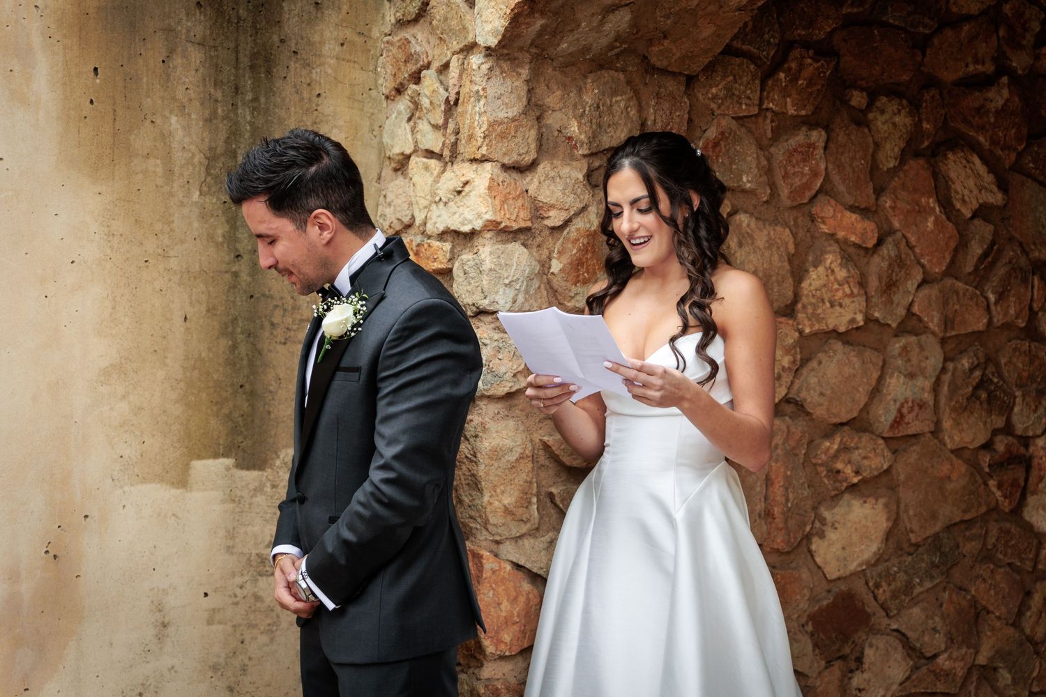 A bride and groom share a romantic moment in front of a rustic stone wall during their wedding ceremony.