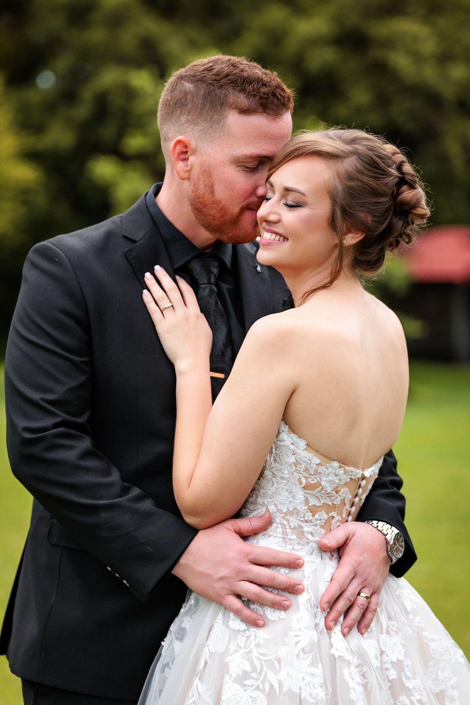 A romantic wedding couple embraces in a garden setting wearing formal attire with a white lace gown and black suit.