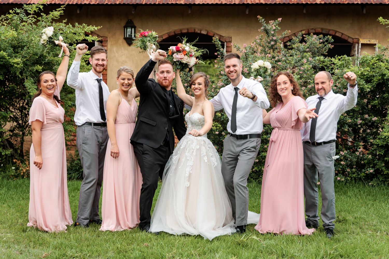 Wedding party in pink bridesmaid dresses and gray suits pose together for formal outdoor photos in a garden setting.