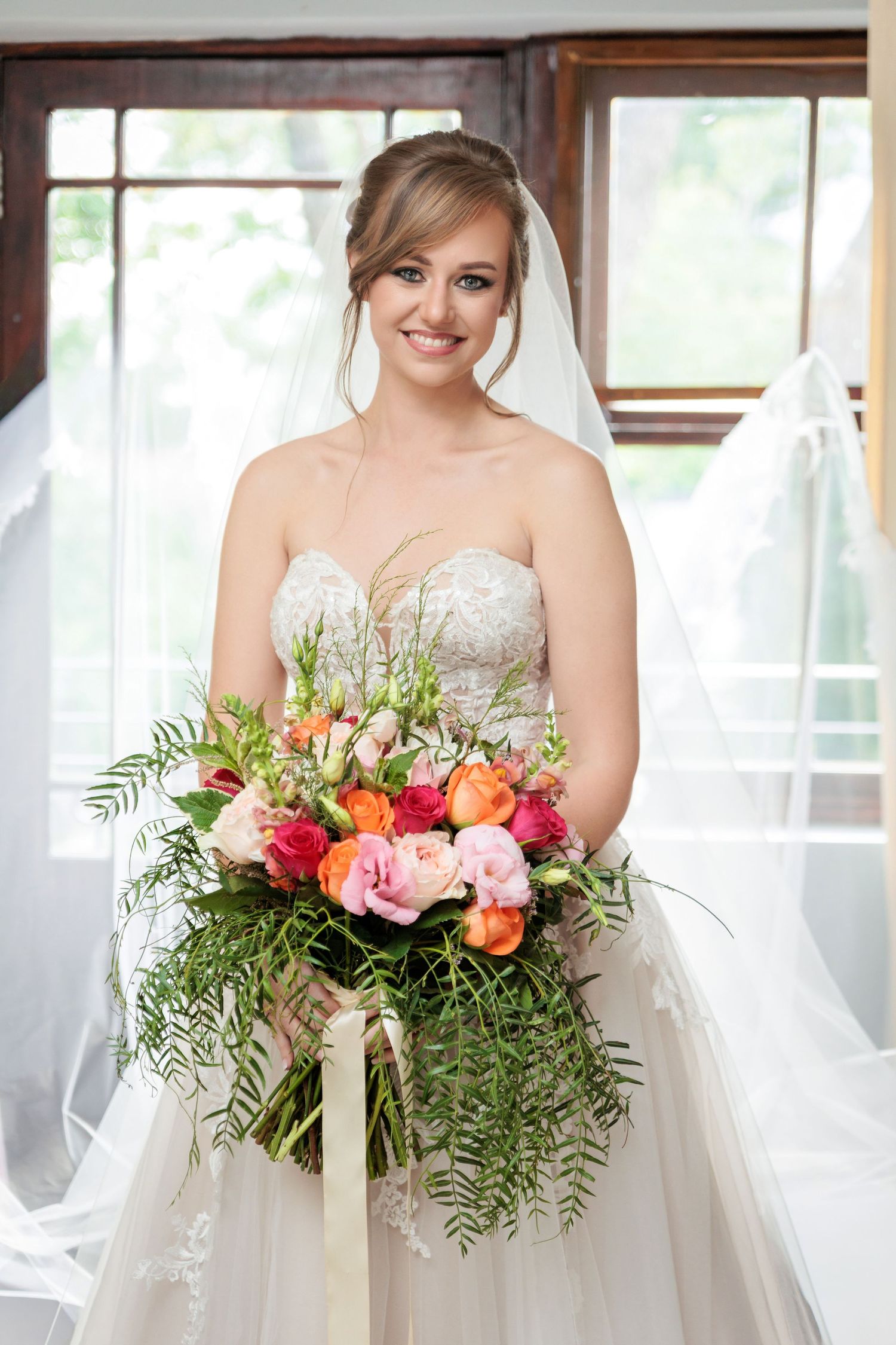 A wedding bouquet with pink and orange flowers and ferns held against a white dress in natural window light.