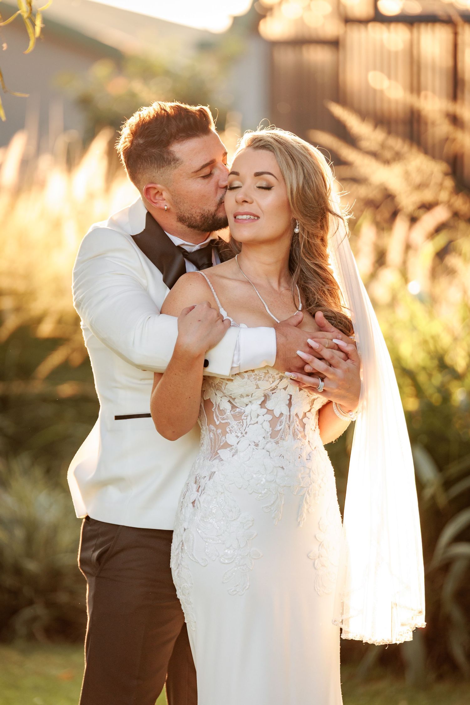 A romantic wedding couple embraces in golden sunlight with tall grass and a veil flowing in the background.
