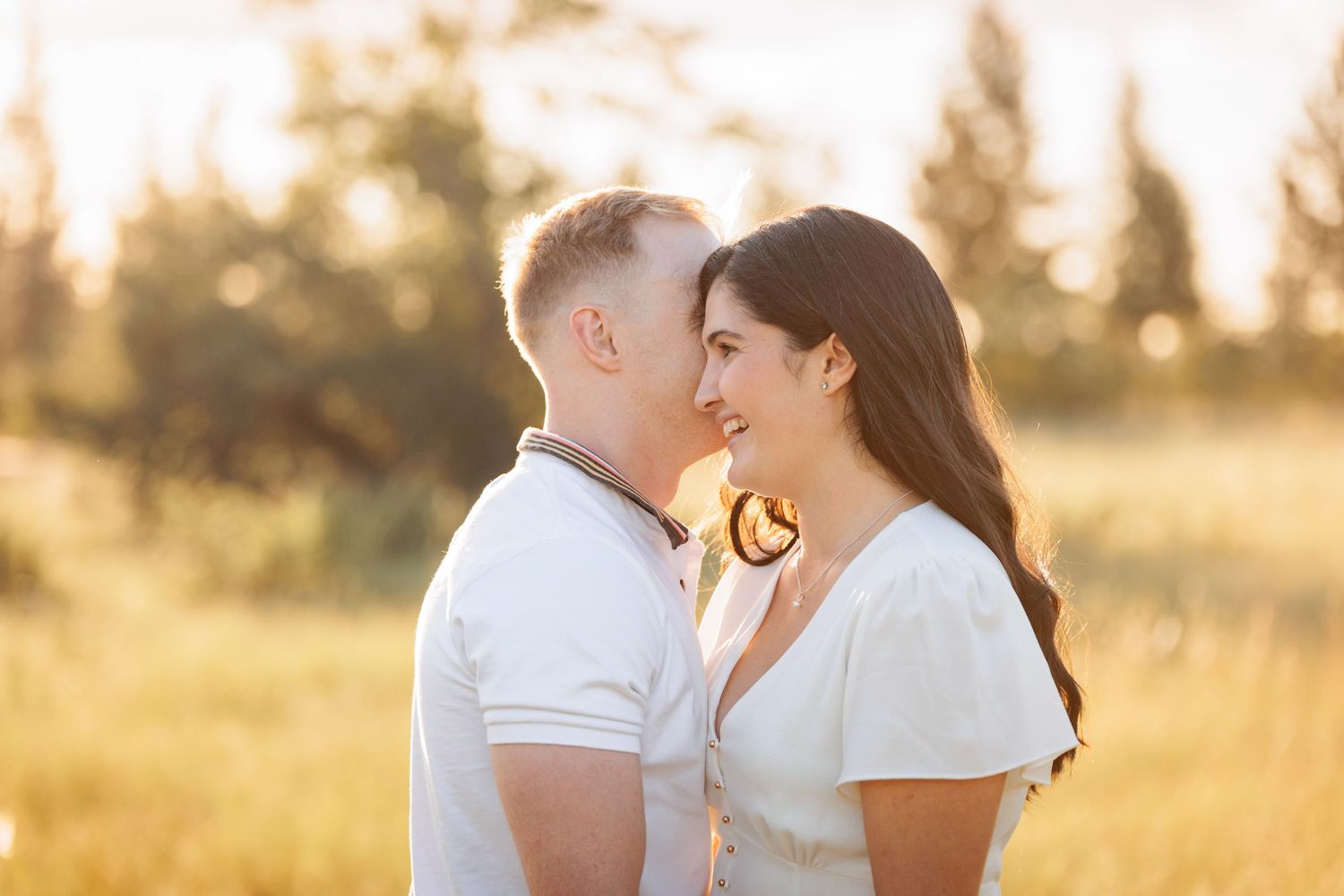 A couple shares a tender moment in a sunlit field during golden hour while wearing white clothing.
