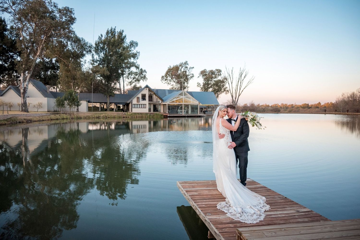 Bride and groom kissing on a wooden dock by a serene lake at sunset with a rustic venue reflected in the water.