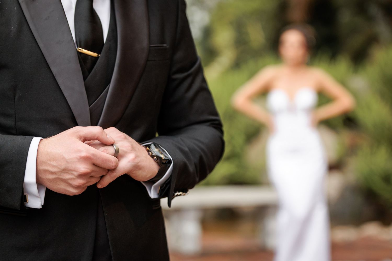A groom adjusts his black suit jacket sleeve while standing outdoors during a wedding ceremony.