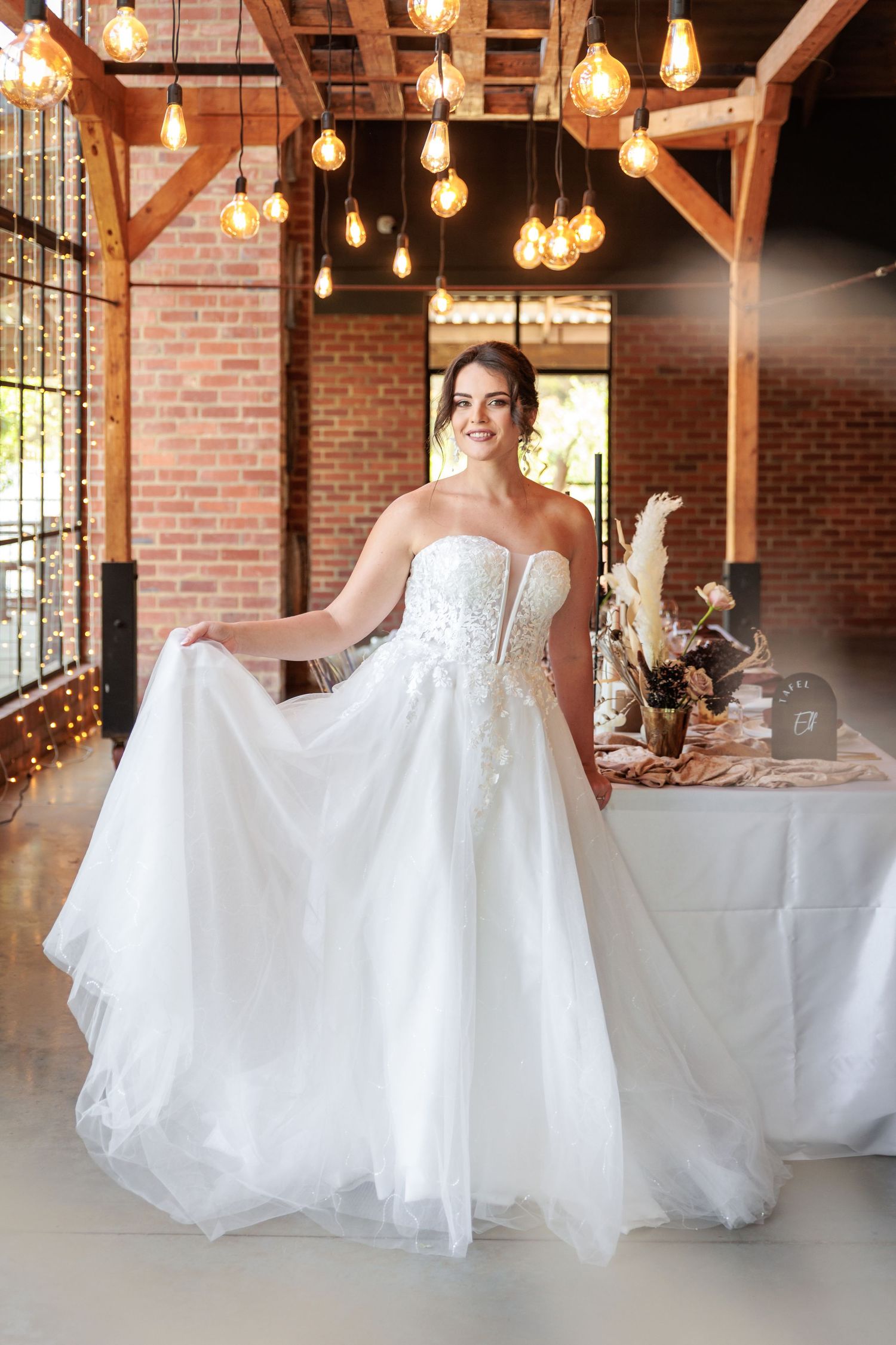 A strapless white wedding gown with tulle skirt is showcased in a rustic brick venue with hanging Edison bulb lighting.