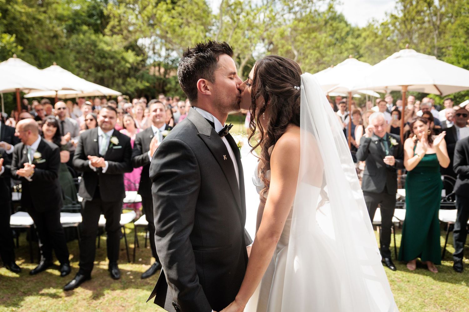 A couple shares their first kiss at an outdoor garden wedding ceremony as guests cheer and applaud in the background.