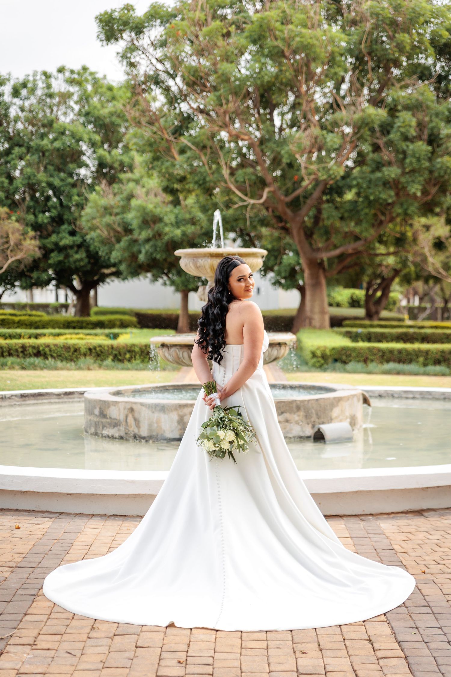 A stunning white wedding dress spreads out in a circular shape on brick pavers surrounded by lush garden scenery.