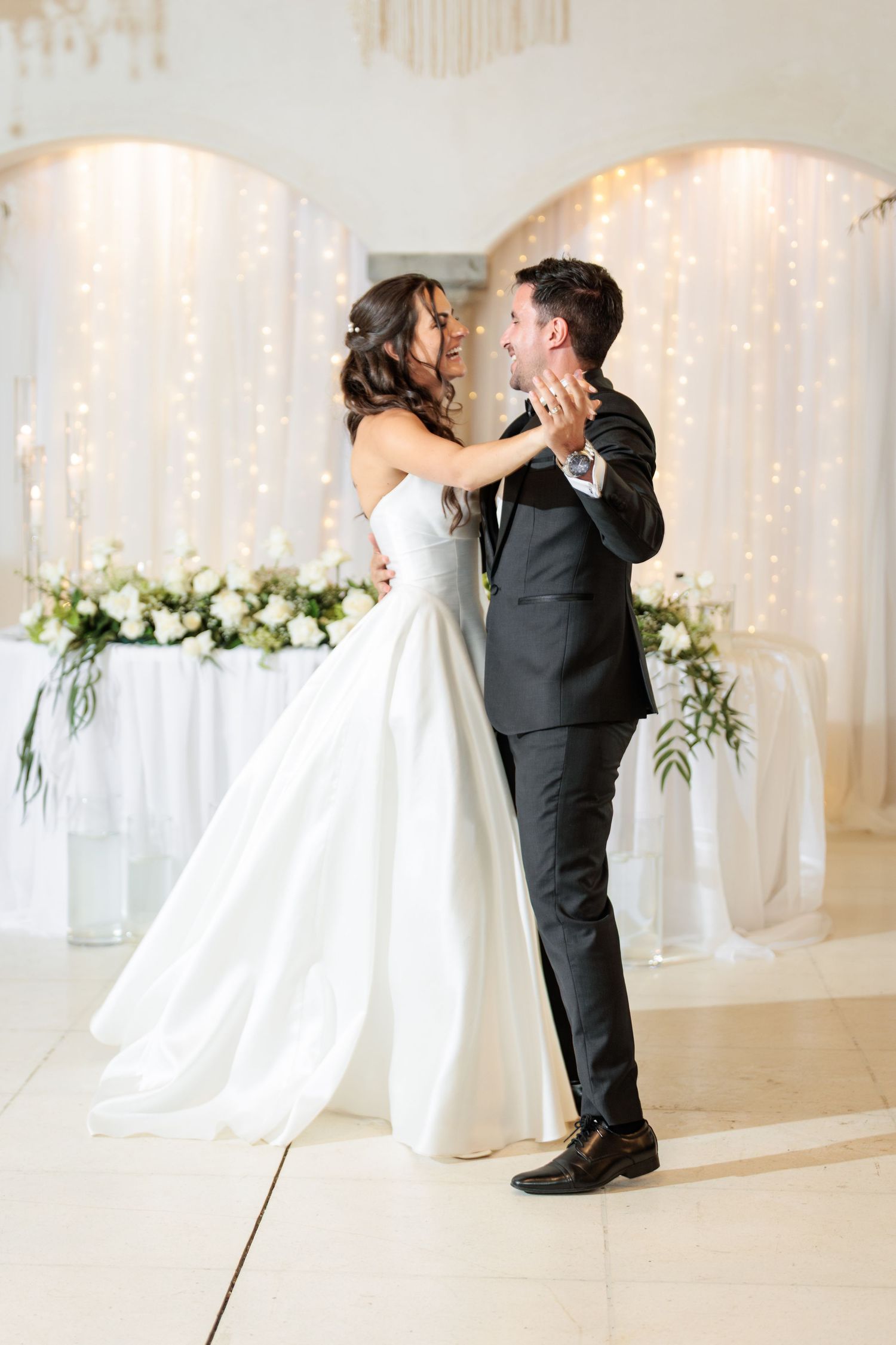 A romantic first dance sequence between newlyweds in an elegant white reception hall with twinkling lights.