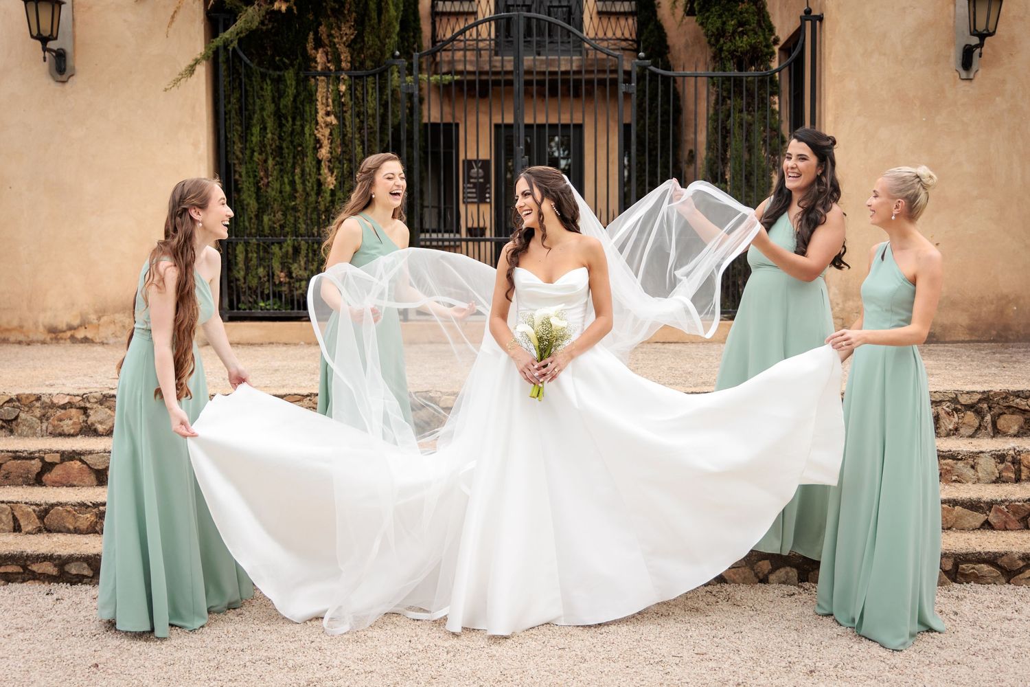 Bridesmaids in mint green dresses help arrange a flowing white wedding dress on stone steps of a Spanish-style building.