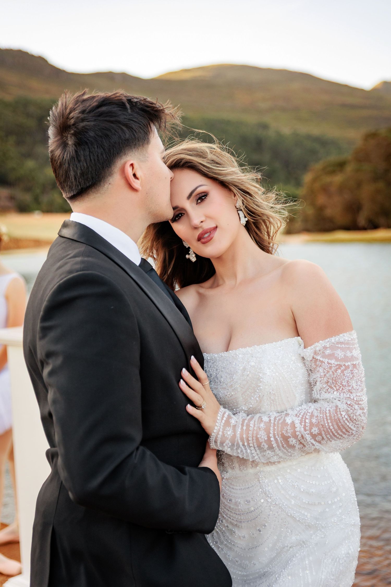 Romantic wedding couple embraces by lakeside at sunset with mountains in background.