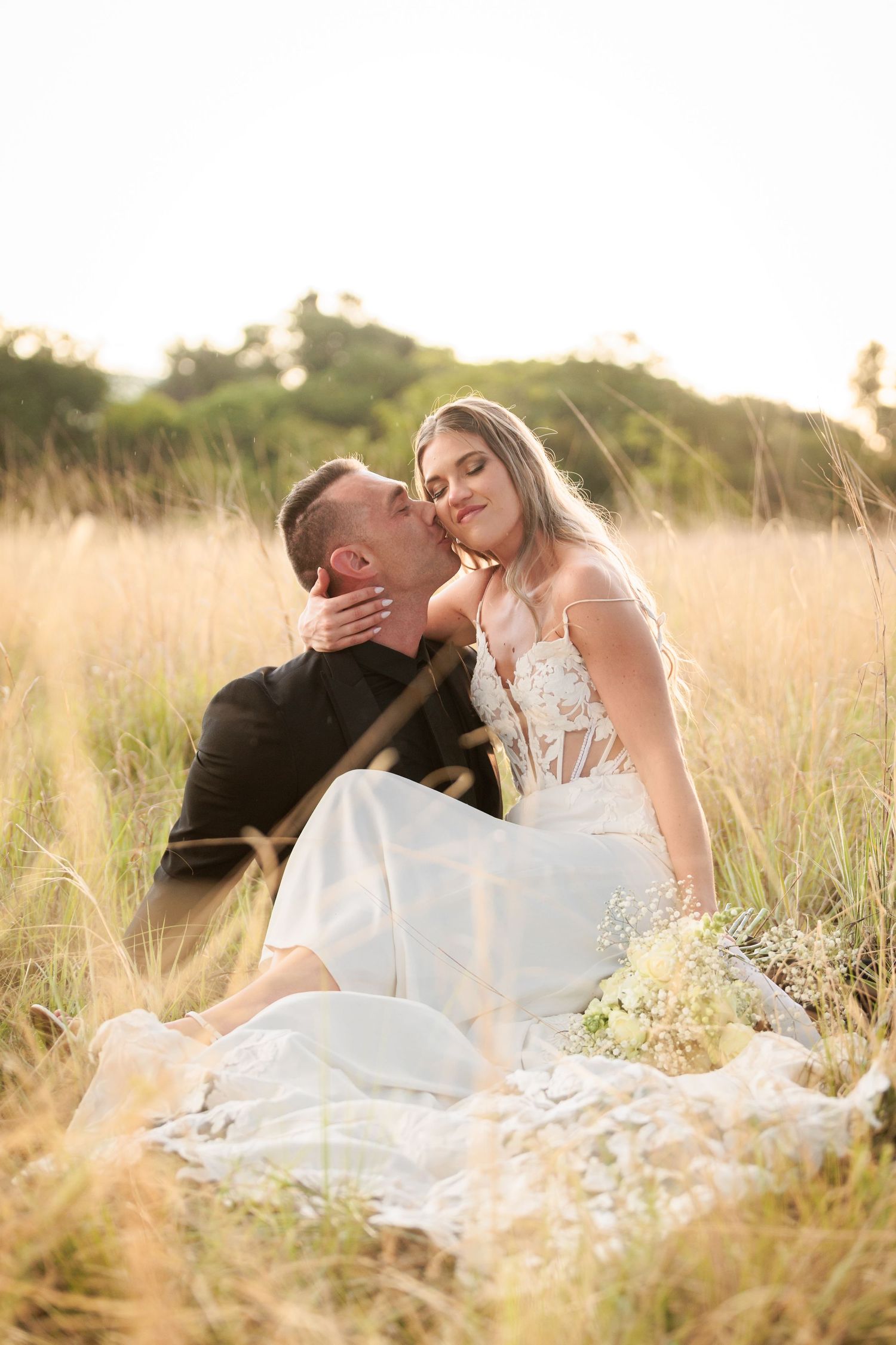A romantic couple in wedding attire embraces while sitting in a sun-lit meadow of tall grass at golden hour.