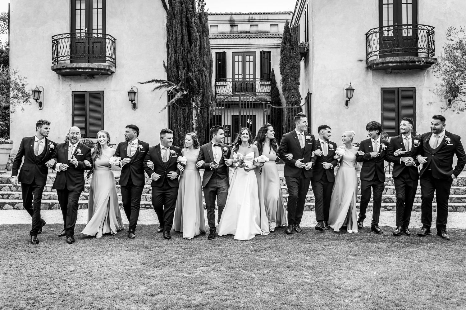 Black and white wedding party photo with bride, groom, and attendants walking arm-in-arm before an elegant villa.