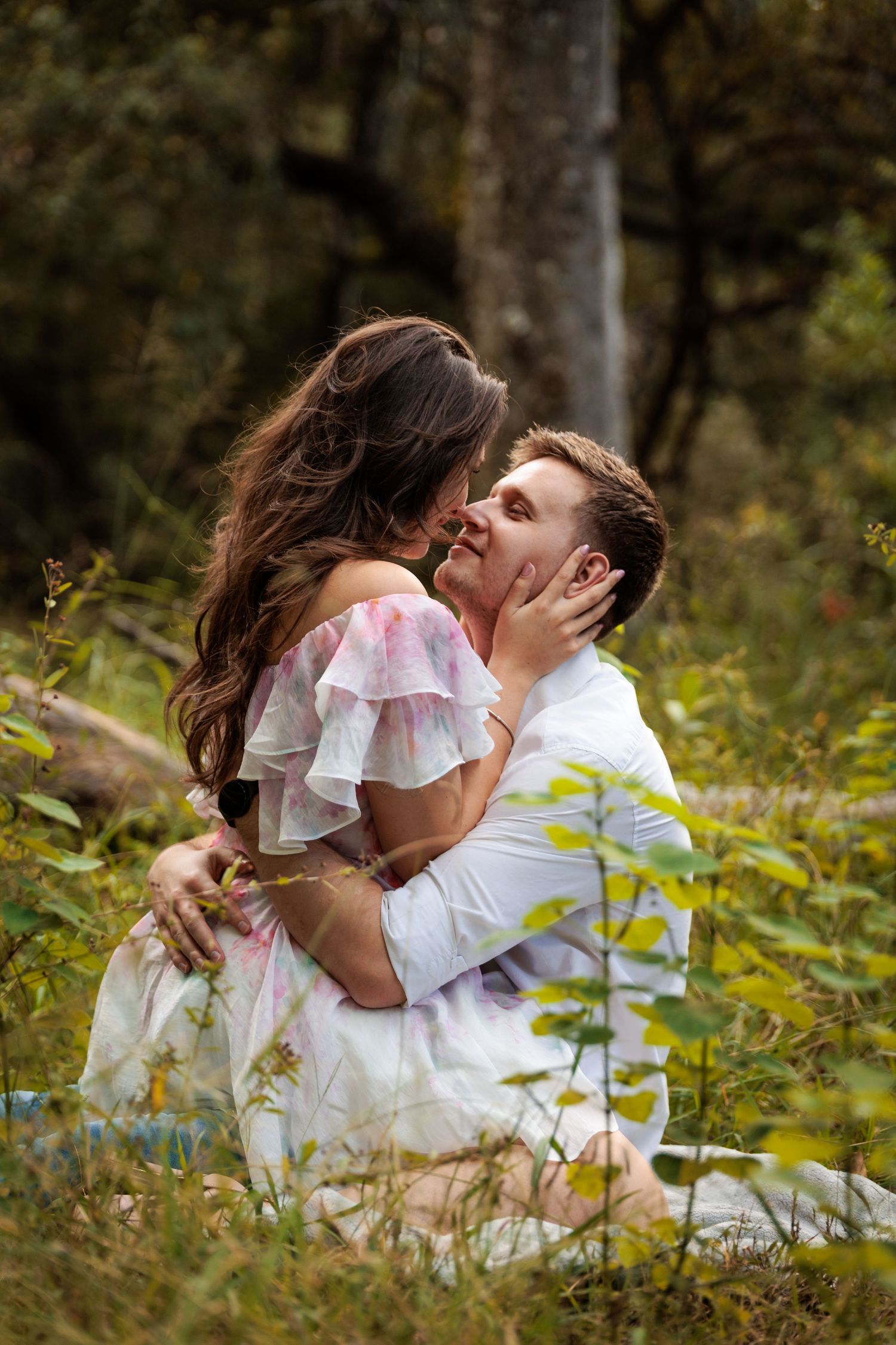 A couple in white outfits sharing a romantic moment in a lush outdoor setting with natural foliage.