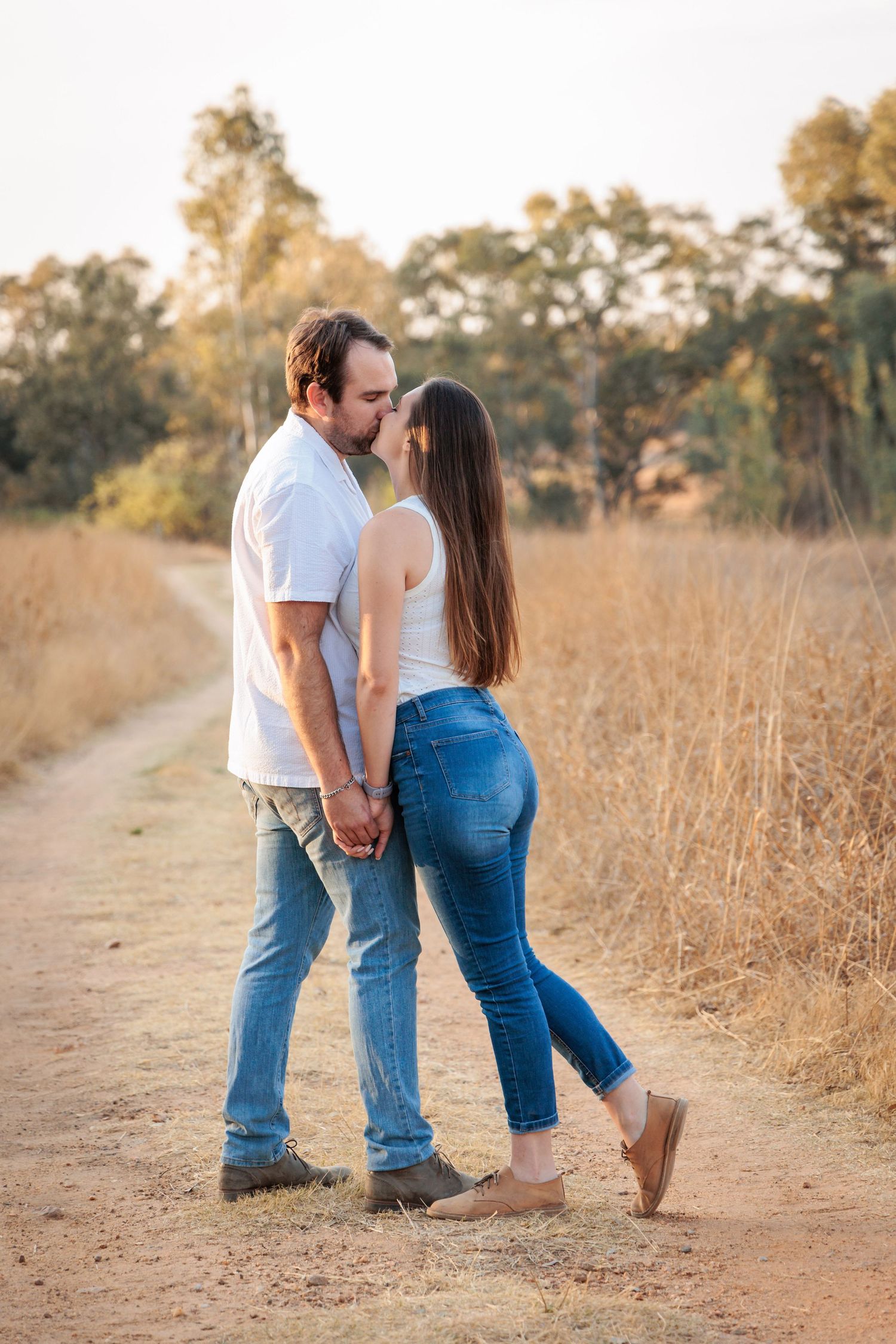 A romantic couple walking on a dirt path through golden grass at sunset wearing matching denim jeans.
