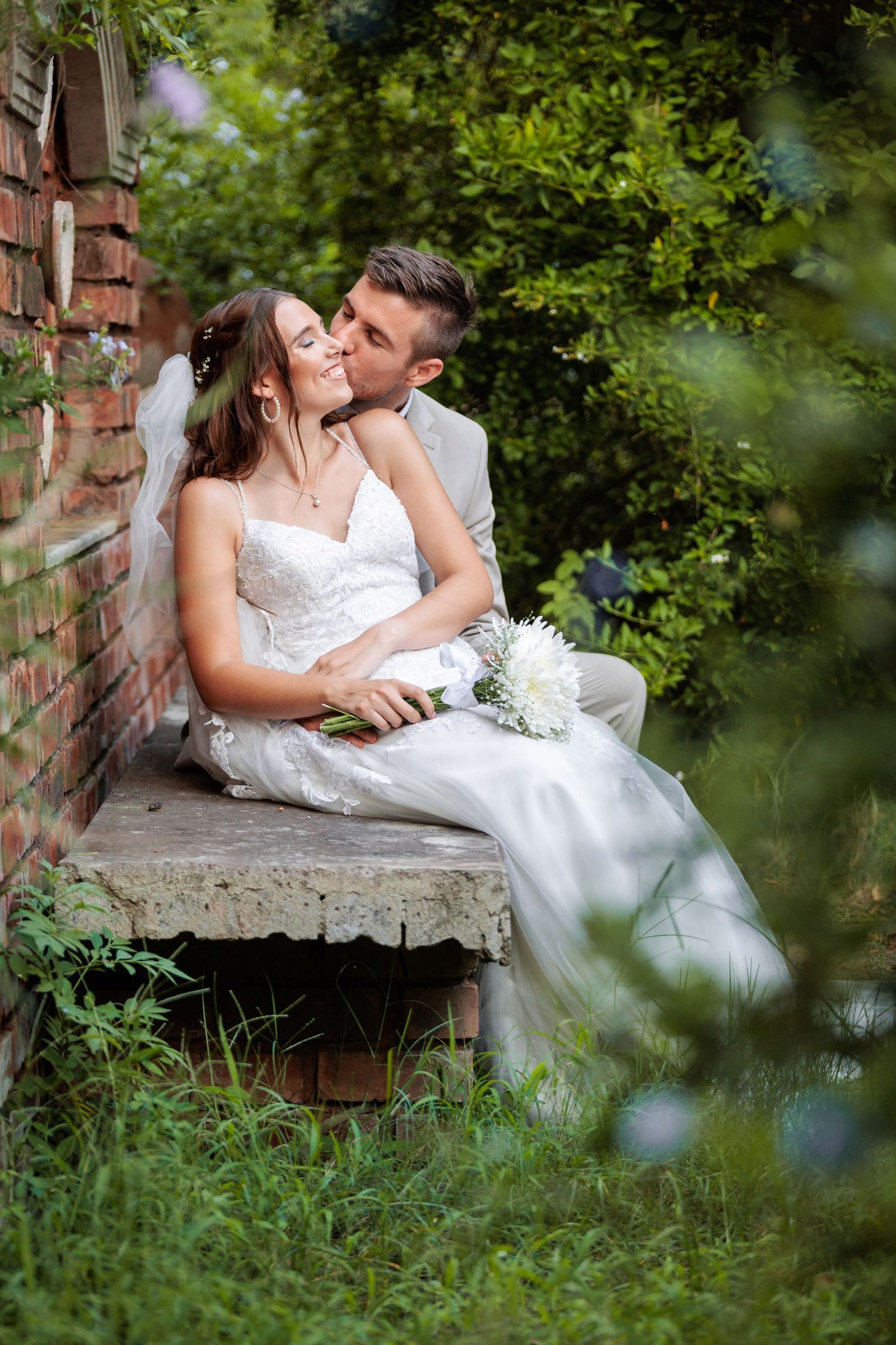 Romantic wedding couple shares intimate moments on a rustic brick bench surrounded by greenery.