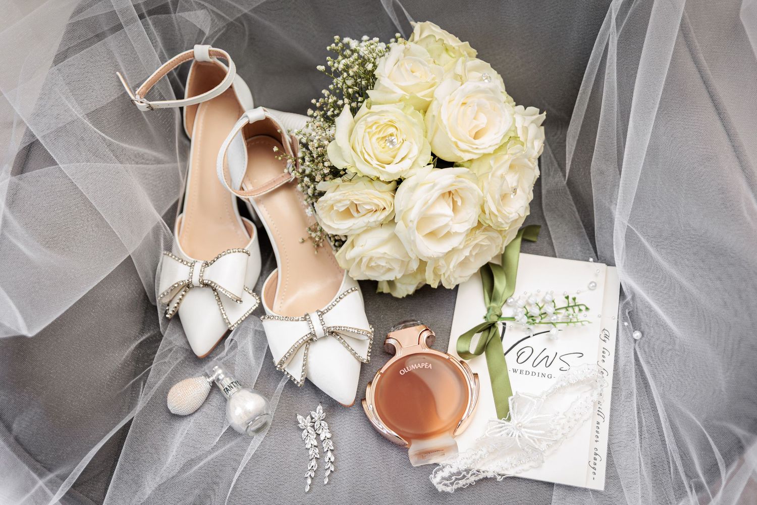 A wedding detail photo showing white heels, tulle veil, and delicate floral bouquet arranged on a light surface.