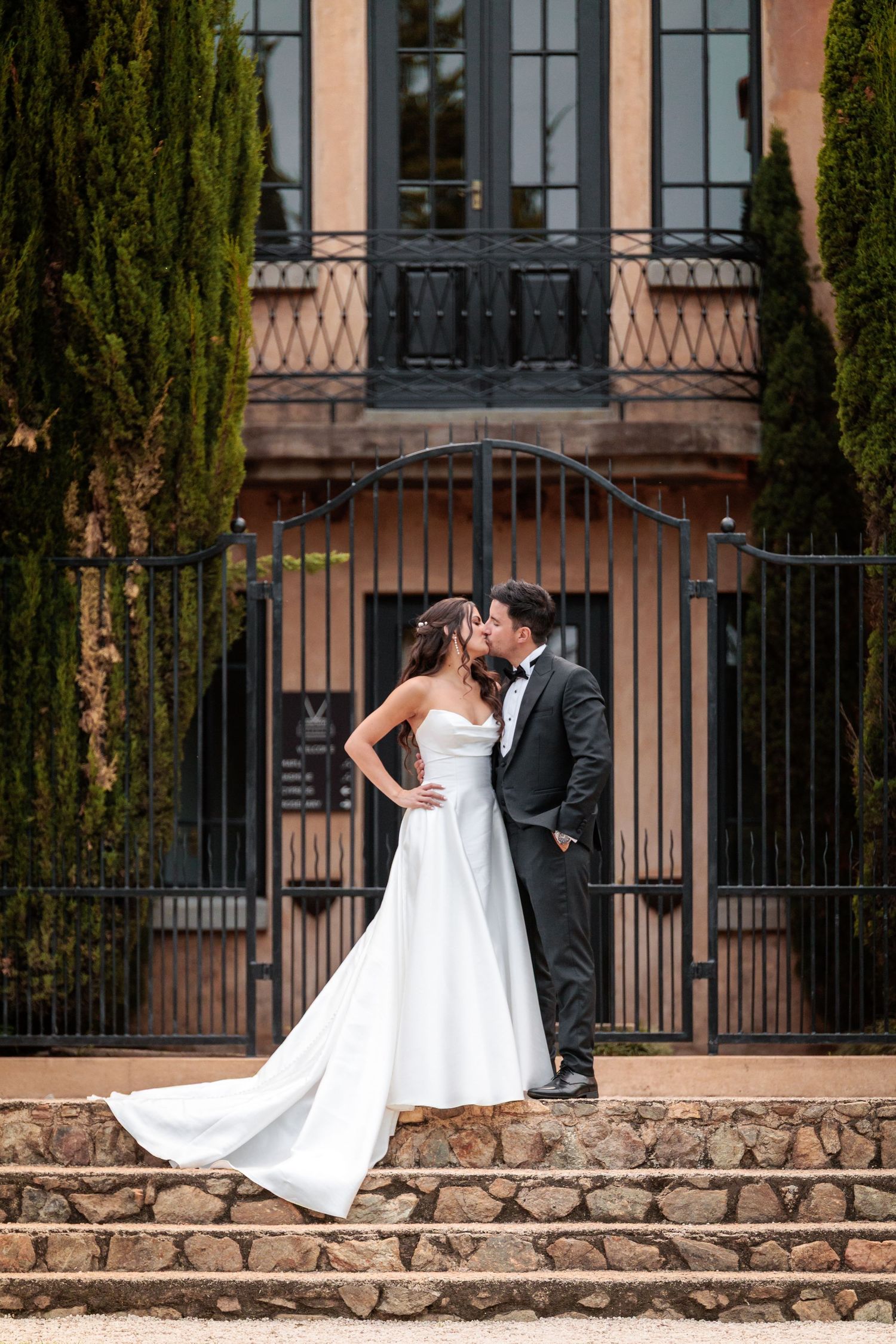 A romantic wedding couple shares an intimate moment on a stone wall with cypress trees and iron gates in the background.