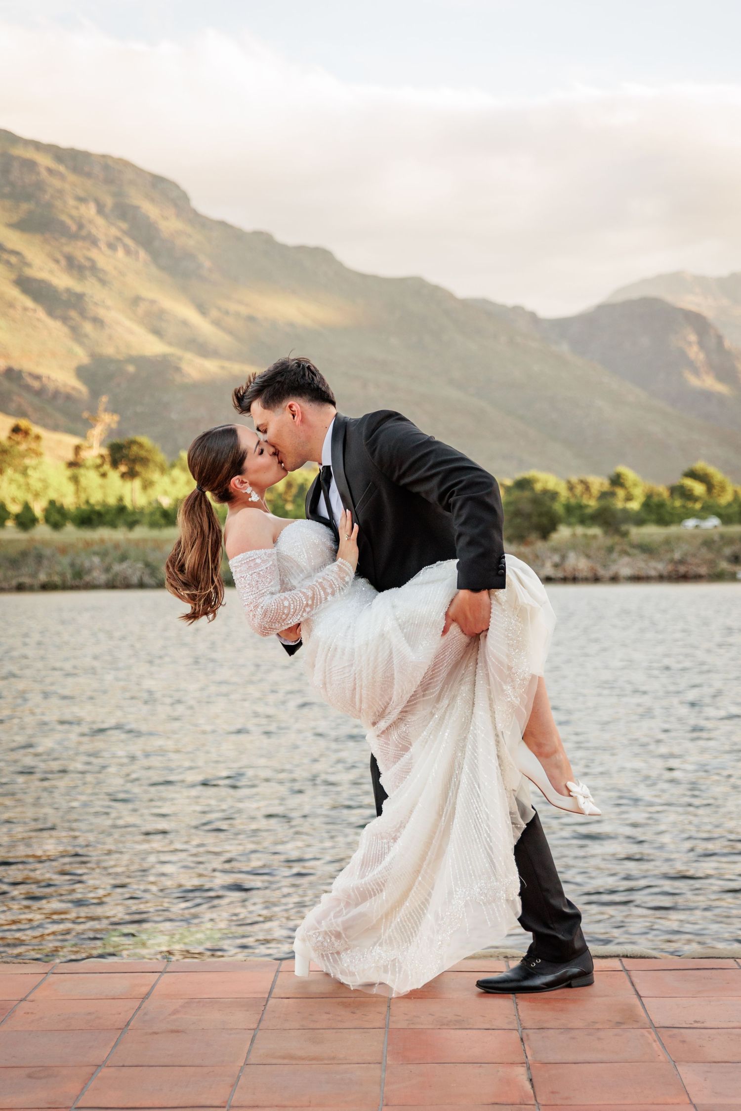 A couple in wedding attire share a romantic dip kiss against a backdrop of mountains and water at sunset.