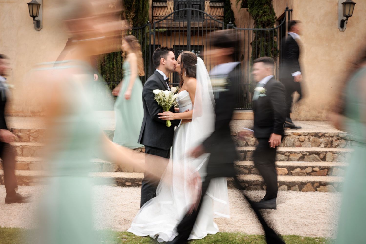 Wedding party walks in a line through a garden courtyard creating a motion blur effect in soft pastel colors.