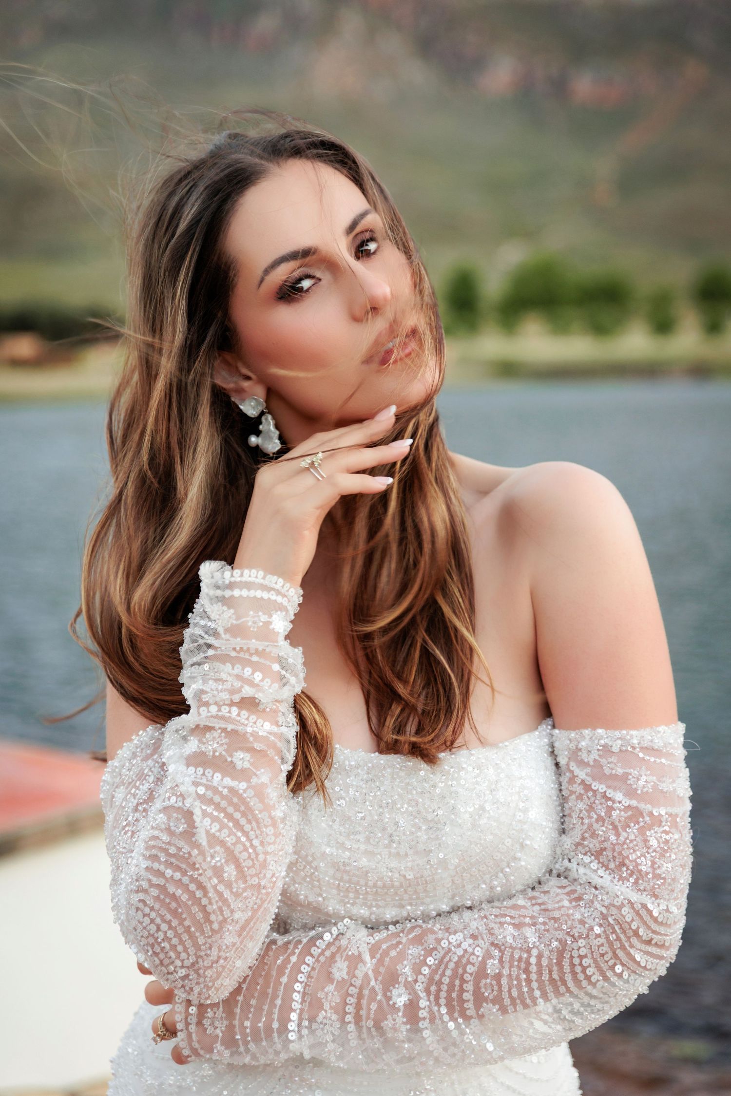 A bride in a white beaded off-shoulder gown with sheer long sleeves poses elegantly by a lake.