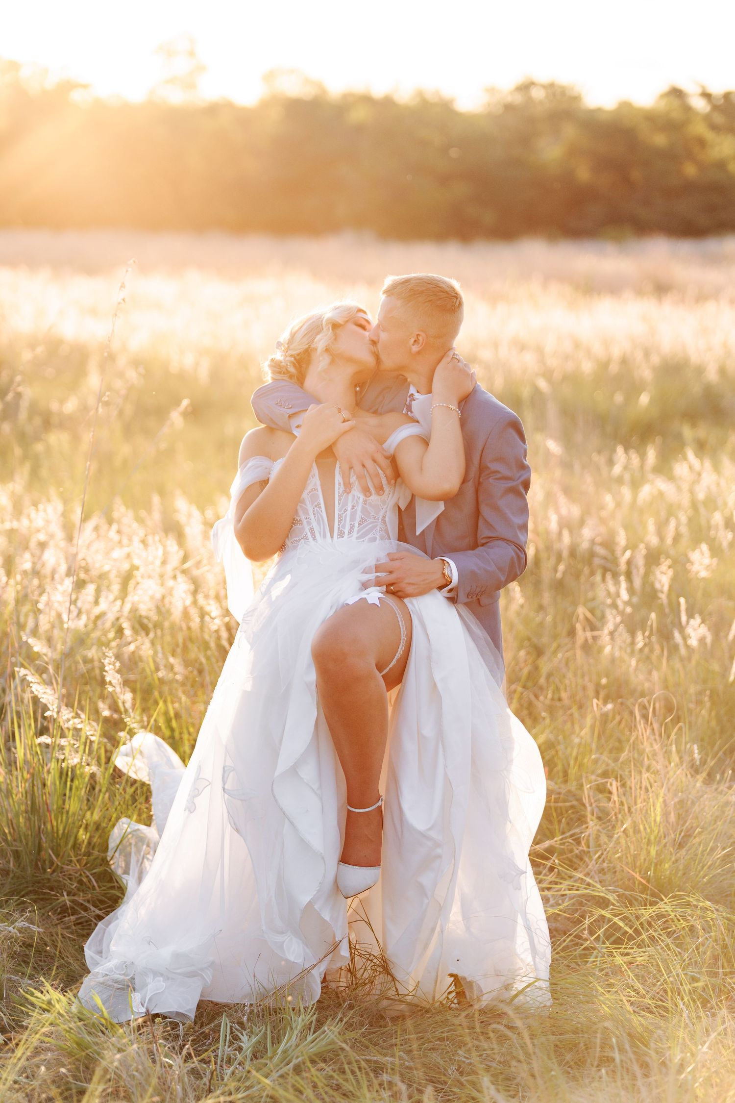 A romantic couple sits together in a sunlit field during golden hour as a white wedding dress flows around them.
