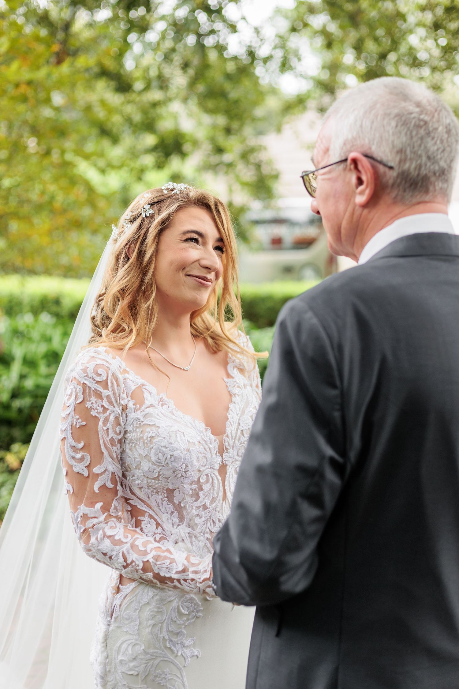 A bride in a lace wedding dress with long sleeves shares a tender moment outdoors with a gentleman in a dark suit.
