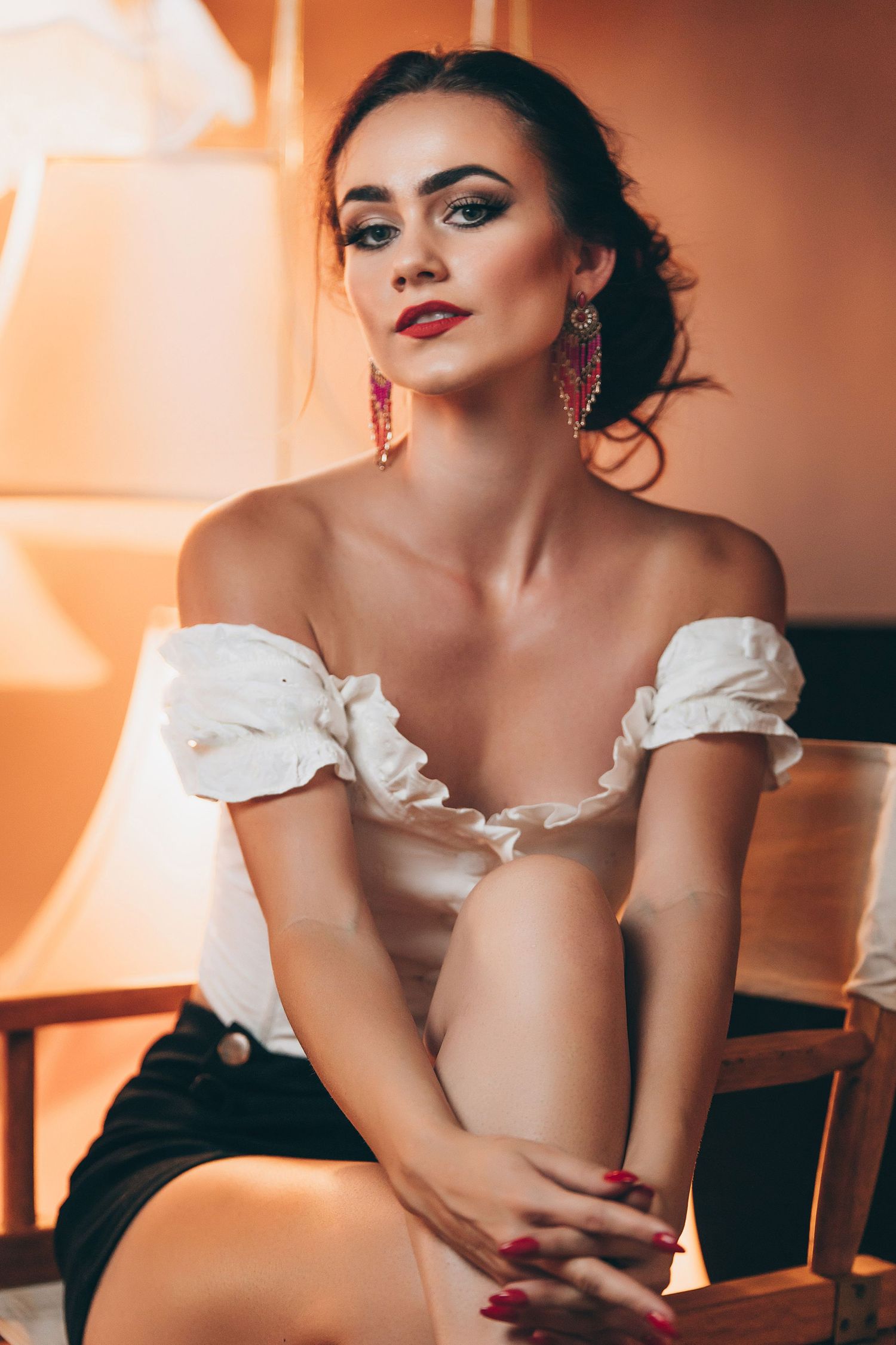 A woman in a white off-shoulder top poses elegantly in warm lighting with dramatic makeup and dark hair.